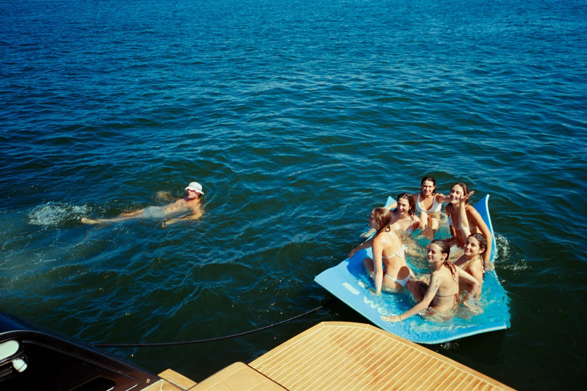 Group of young people on a blue inflatable raft enjoying a day on the water, with some swimming nearby, under a clear sky.