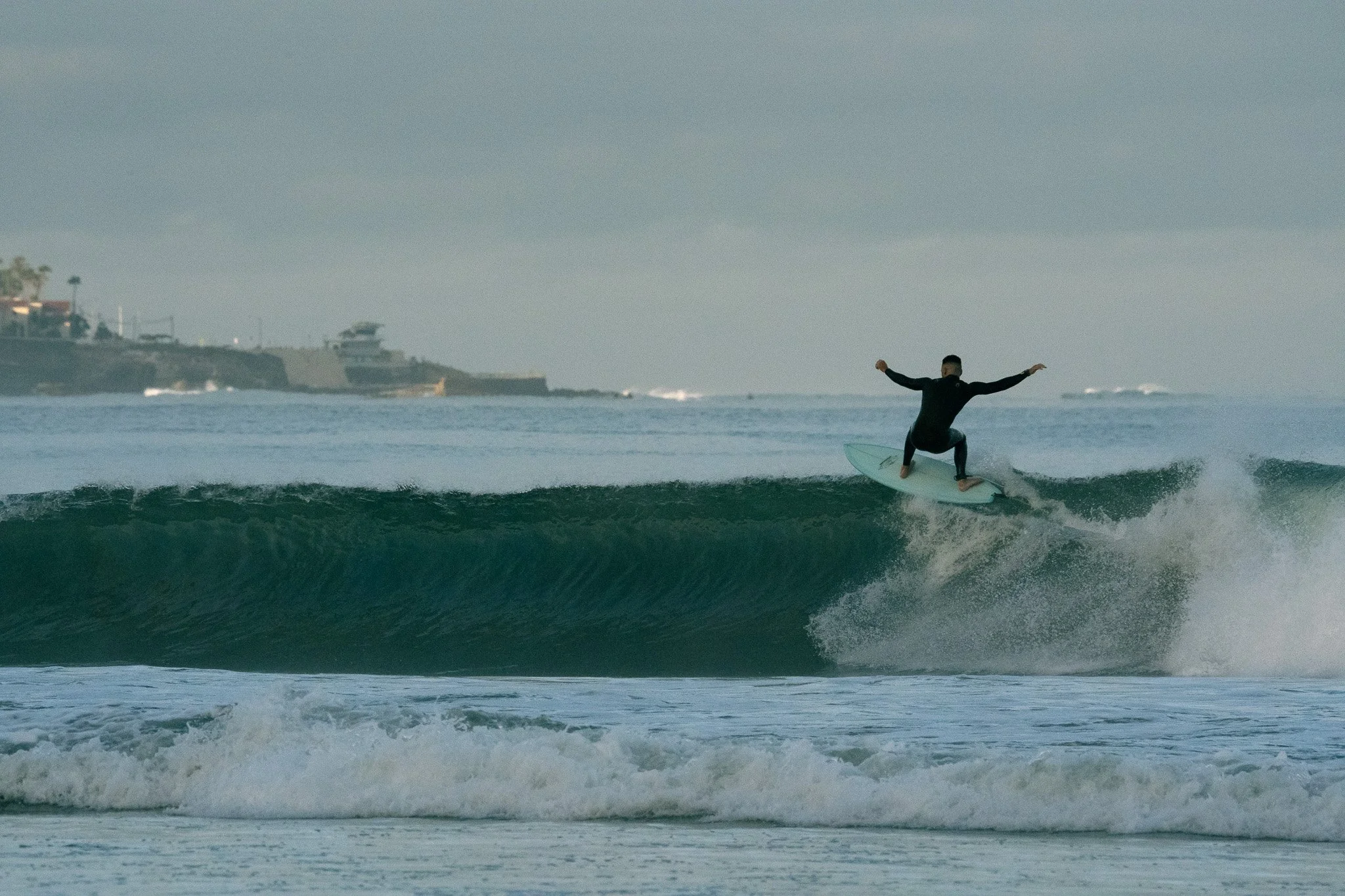 A person surfing on a wave in the ocean with a distant shoreline and buildings in the background.