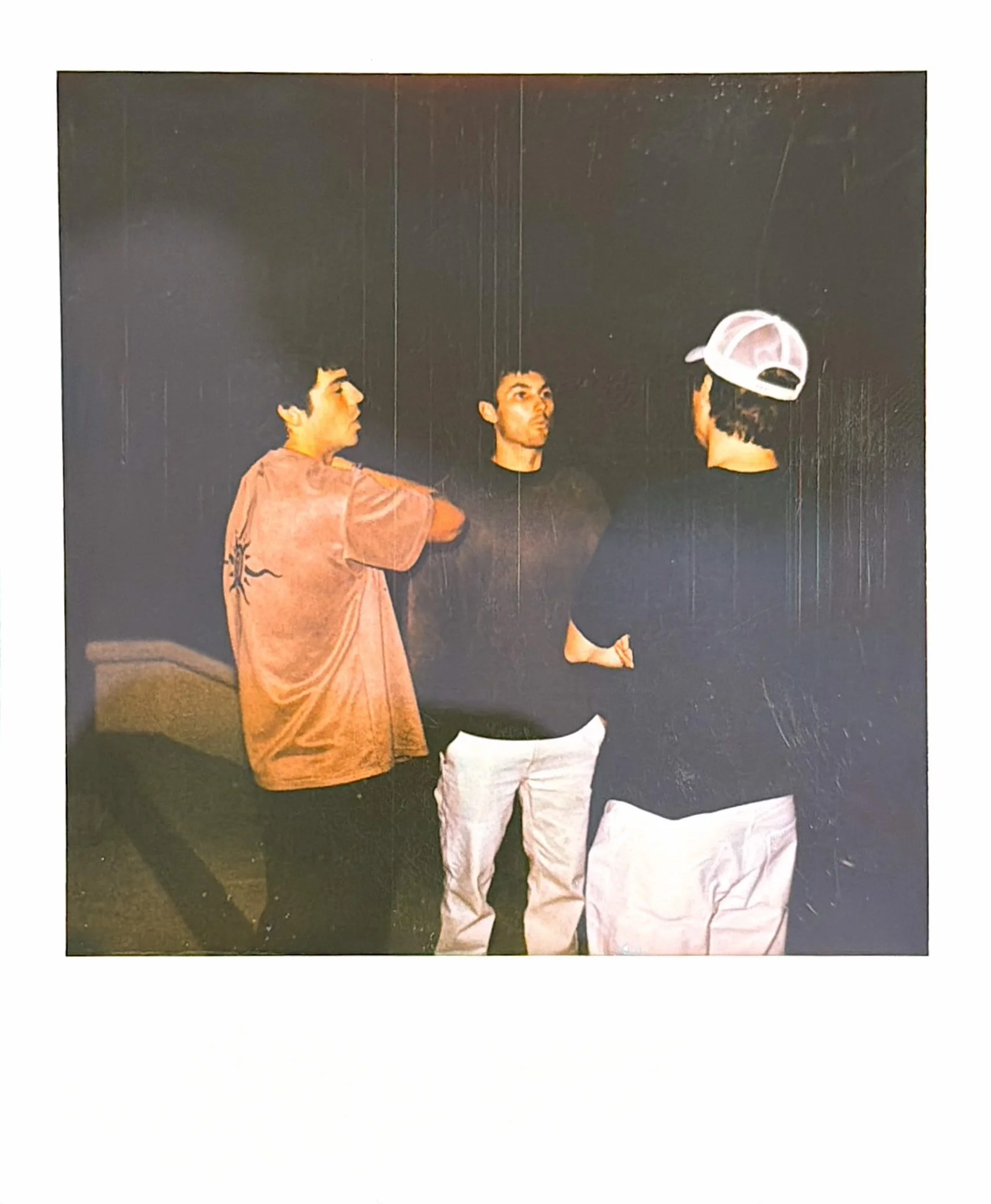 Three men, stand against a black background. The man on the left is wearing a short-sleeved shirt with a design on the back, the man in the middle is facing away with a baseball cap, and the man on the right is looking towards the man.