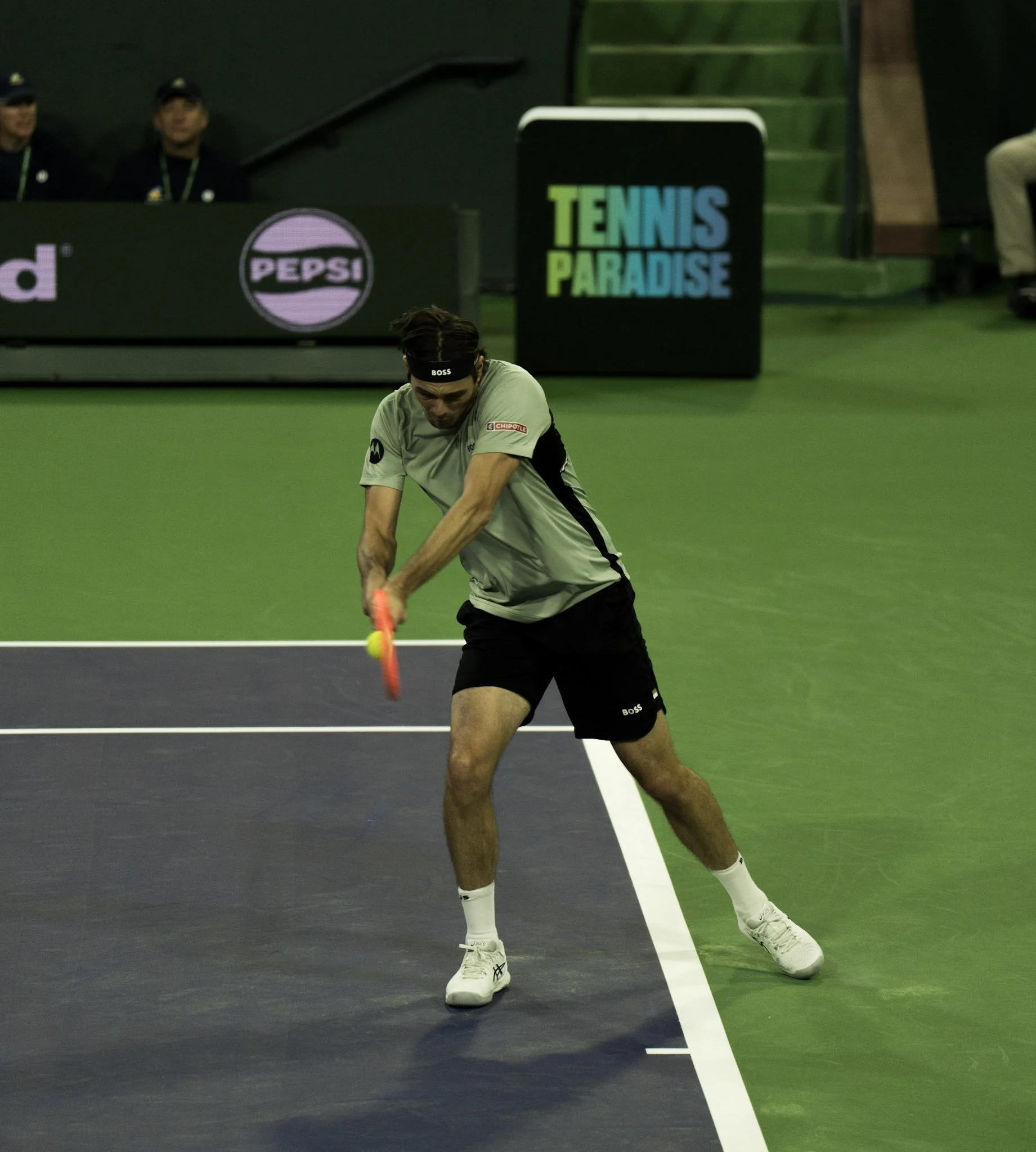 A male tennis player in a grey shirt and black shorts practicing a shot on an indoor tennis court. He is wearing white sneakers and a black headband with the word "BOSS" on it. The background includes a green staircase and a black sign with colorful lettering that says "TENNIS PARADISE", and a black banner with the Pepsi logo. There are three individuals sitting in the background, one wearing a black hat.
