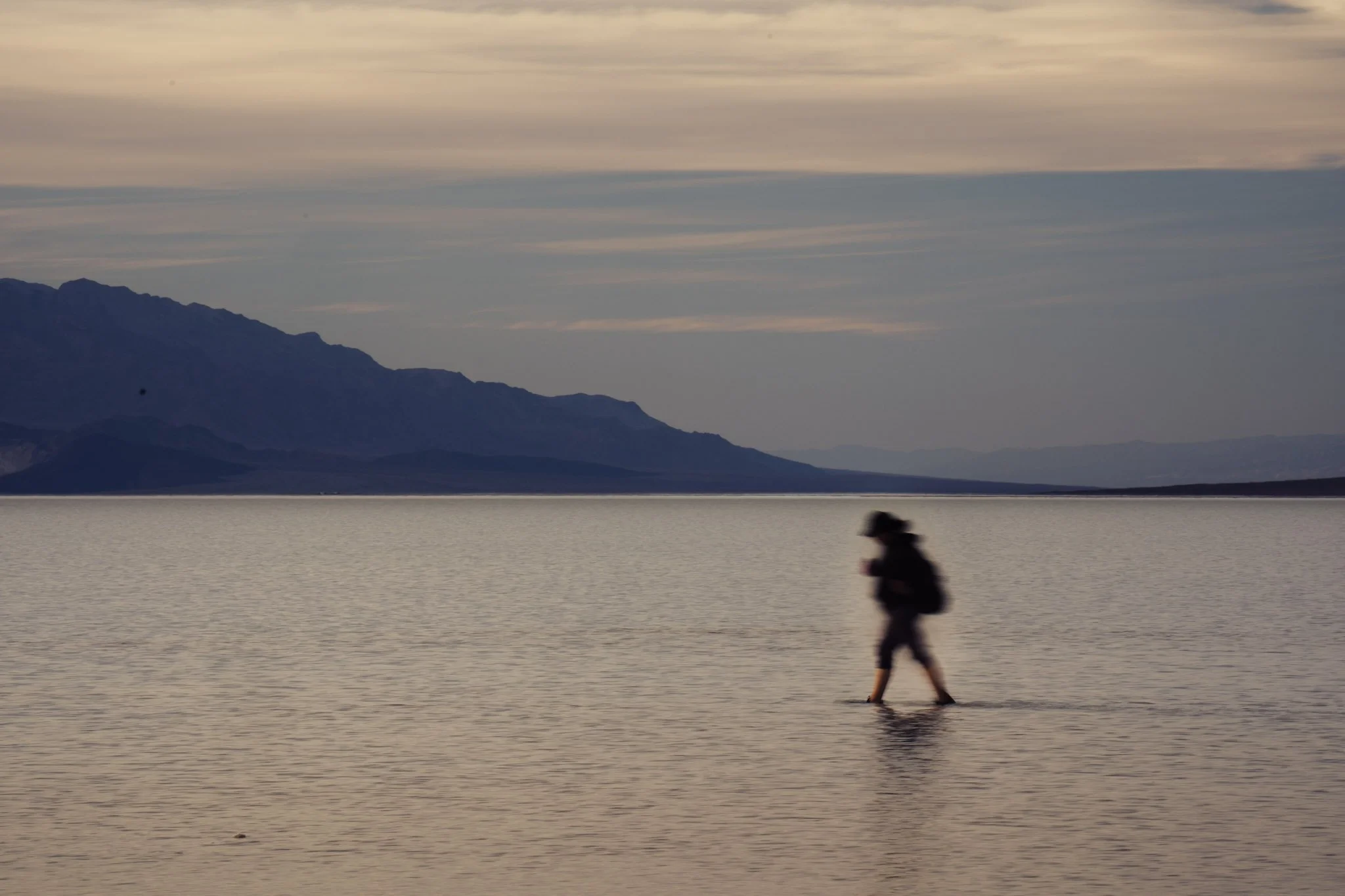 A person walking along the edge of a body of water with mountains in the background under a cloudy sky.