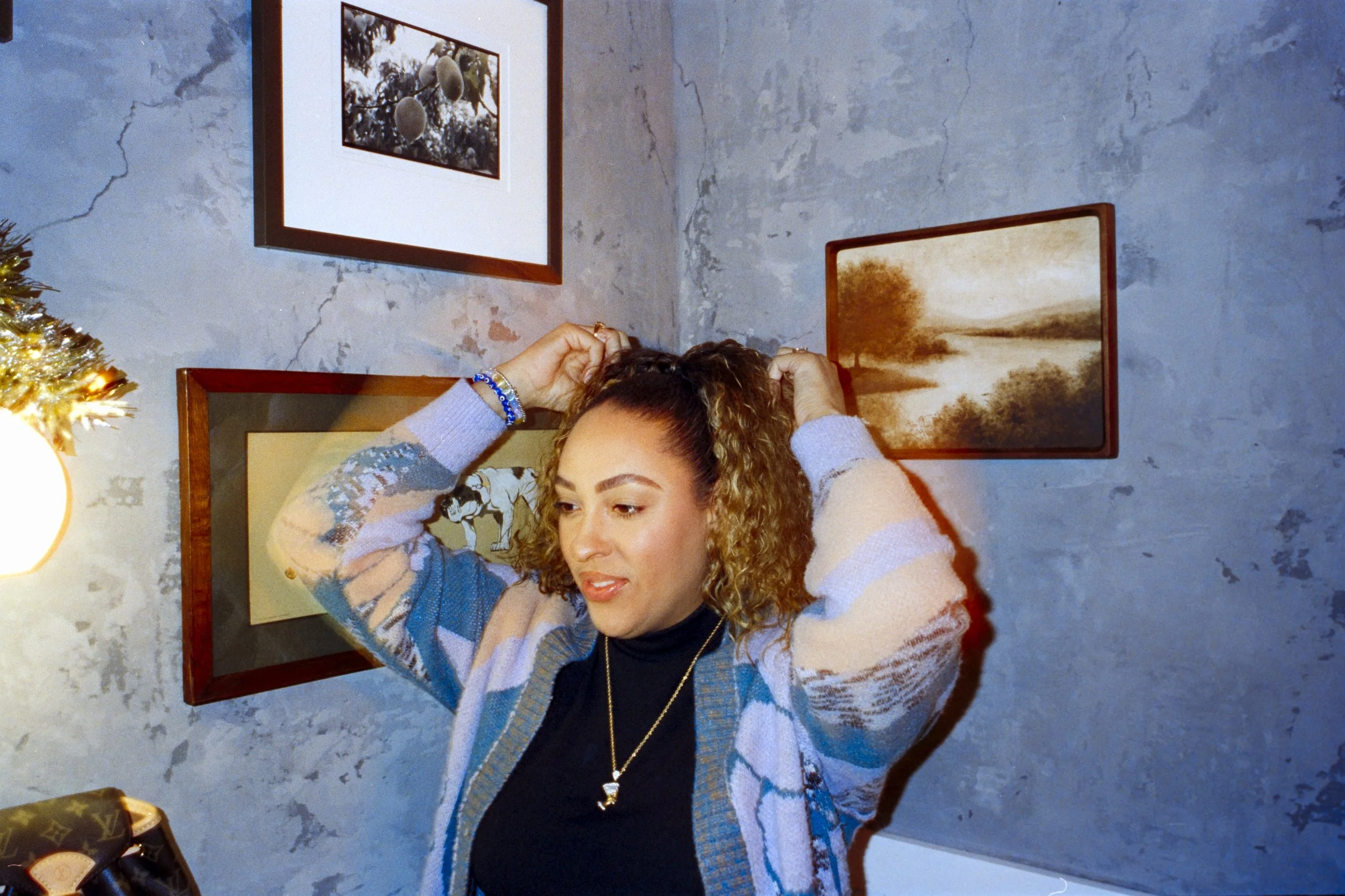 A woman with curly hair adjusts her hair in front of a gray textured wall with framed artwork.