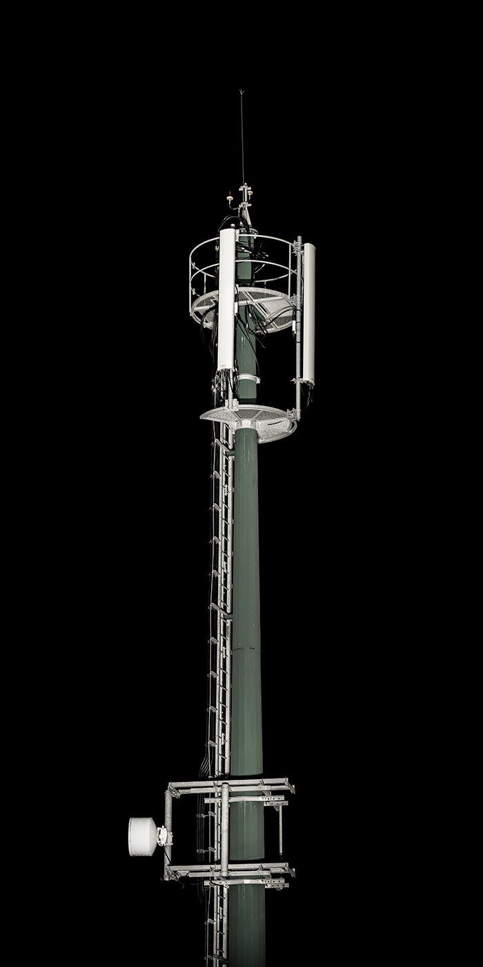 A tall cell phone or radio tower stands against a black night sky, equipped with antennas and safety platforms. SA 02 001, part of the series Allochthonous Systems / Sistemas Alóctones by Miguel Cruz de Carvalho