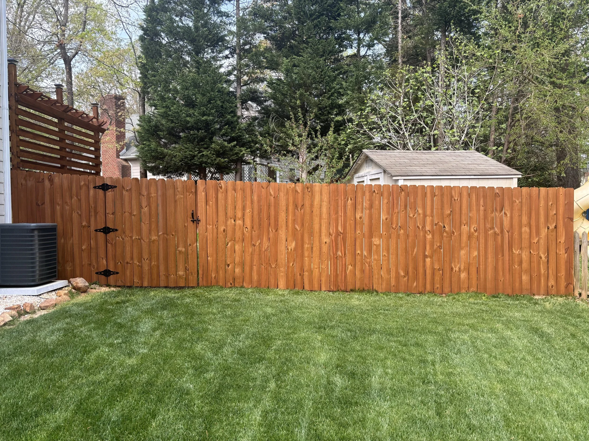 A backyard scene with a green lawn, a wooden fence, and tall trees in the background. There is a small white shed, a section of brick house, and a gray air conditioning unit on the left. The sky is partly cloudy.