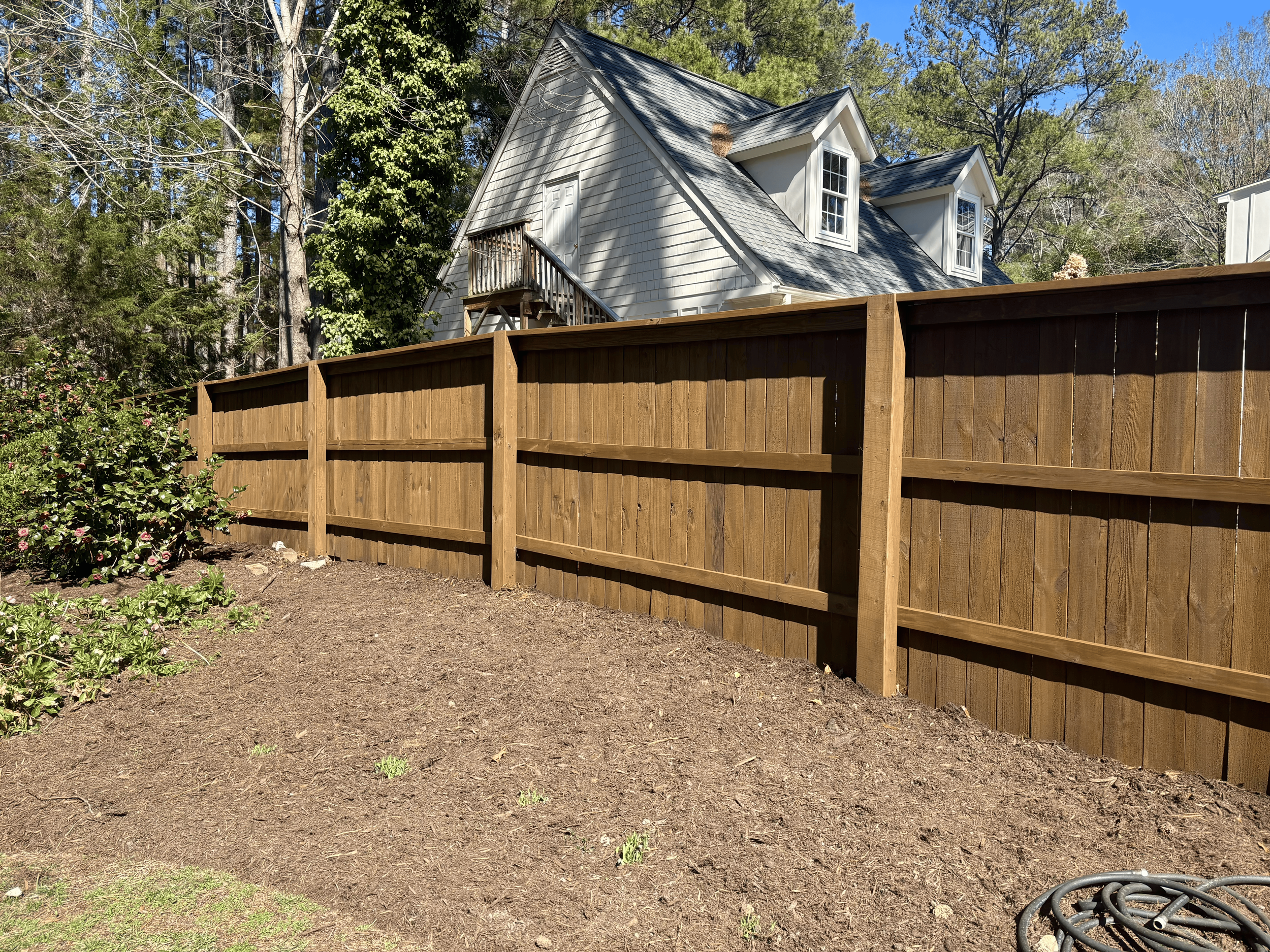 A backyard with a newly built wooden fence, a dirt ground, and a white house with a steep roof and multiple windows, surrounded by trees and greenery.