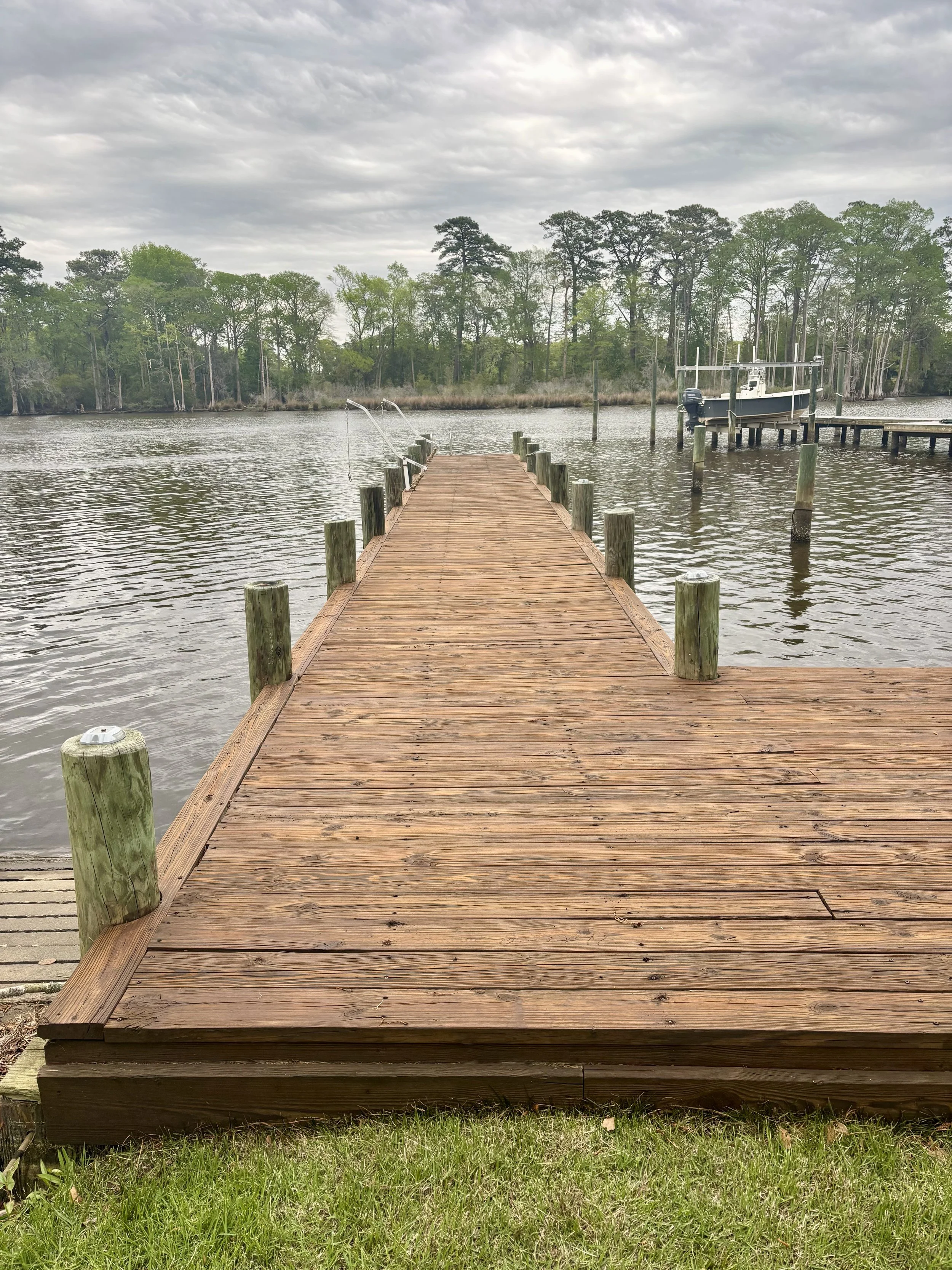 A wooden dock extending over a body of water with a boat on a lift at the end, surrounded by trees and cloudy sky.