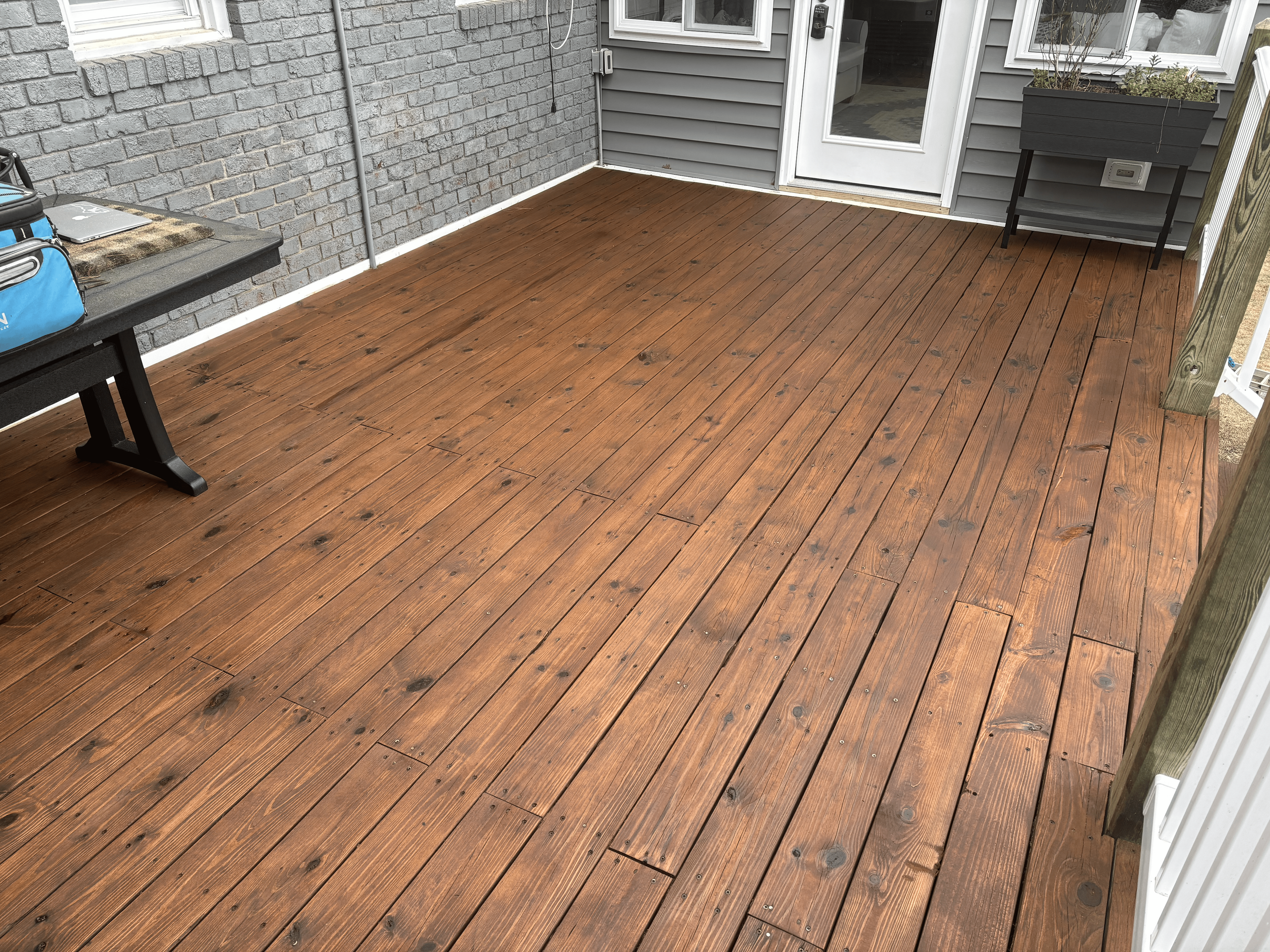A deck with wooden floorboards outside a house with gray siding and a gray brick wall, with a black chair, a small table, and a planter with plants.