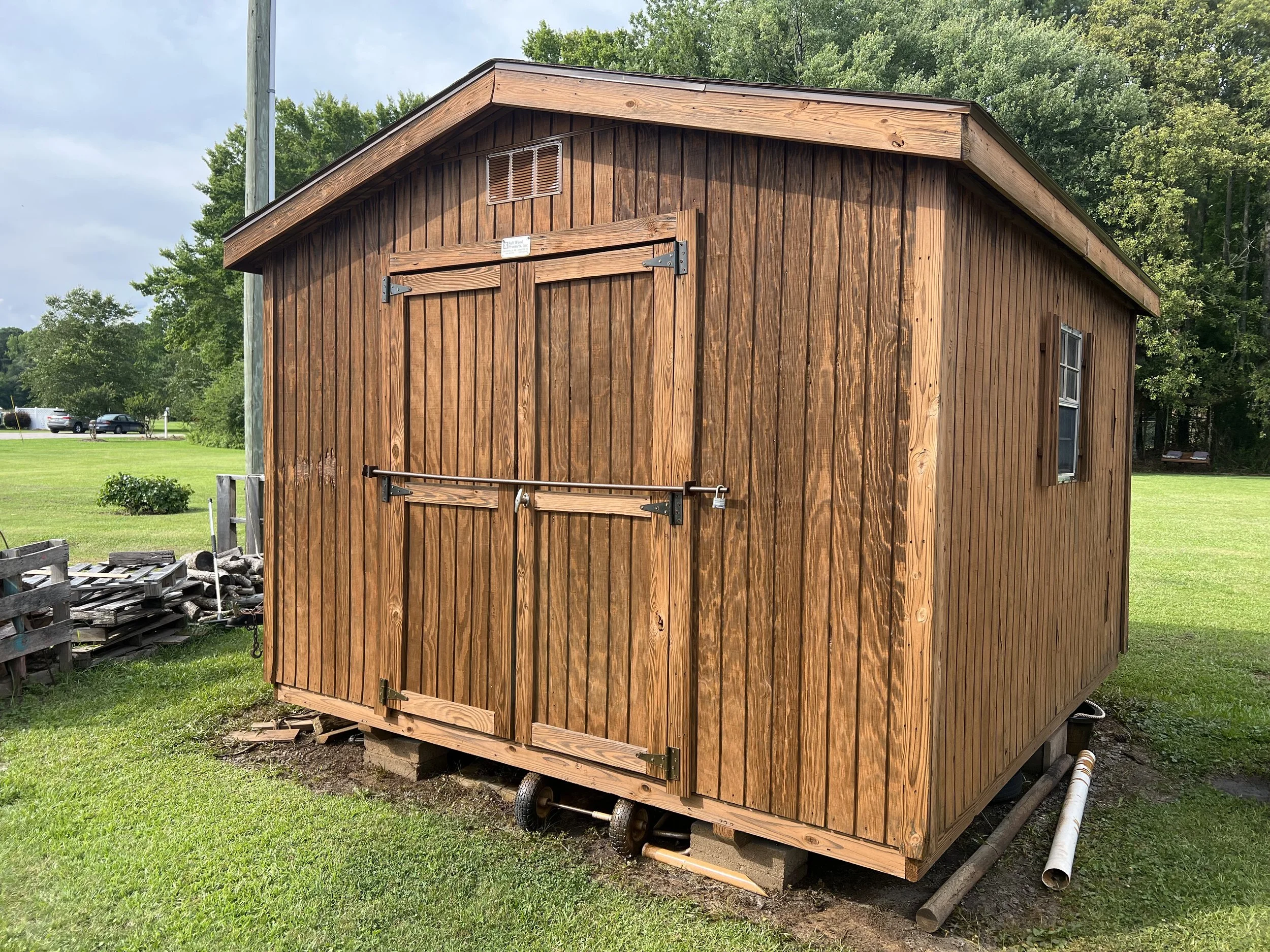 A small wooden shed with a sloped roof, falling apart, with a closed door secured by a padlock, situated on concrete blocks and surrounded by green grass and trees.
