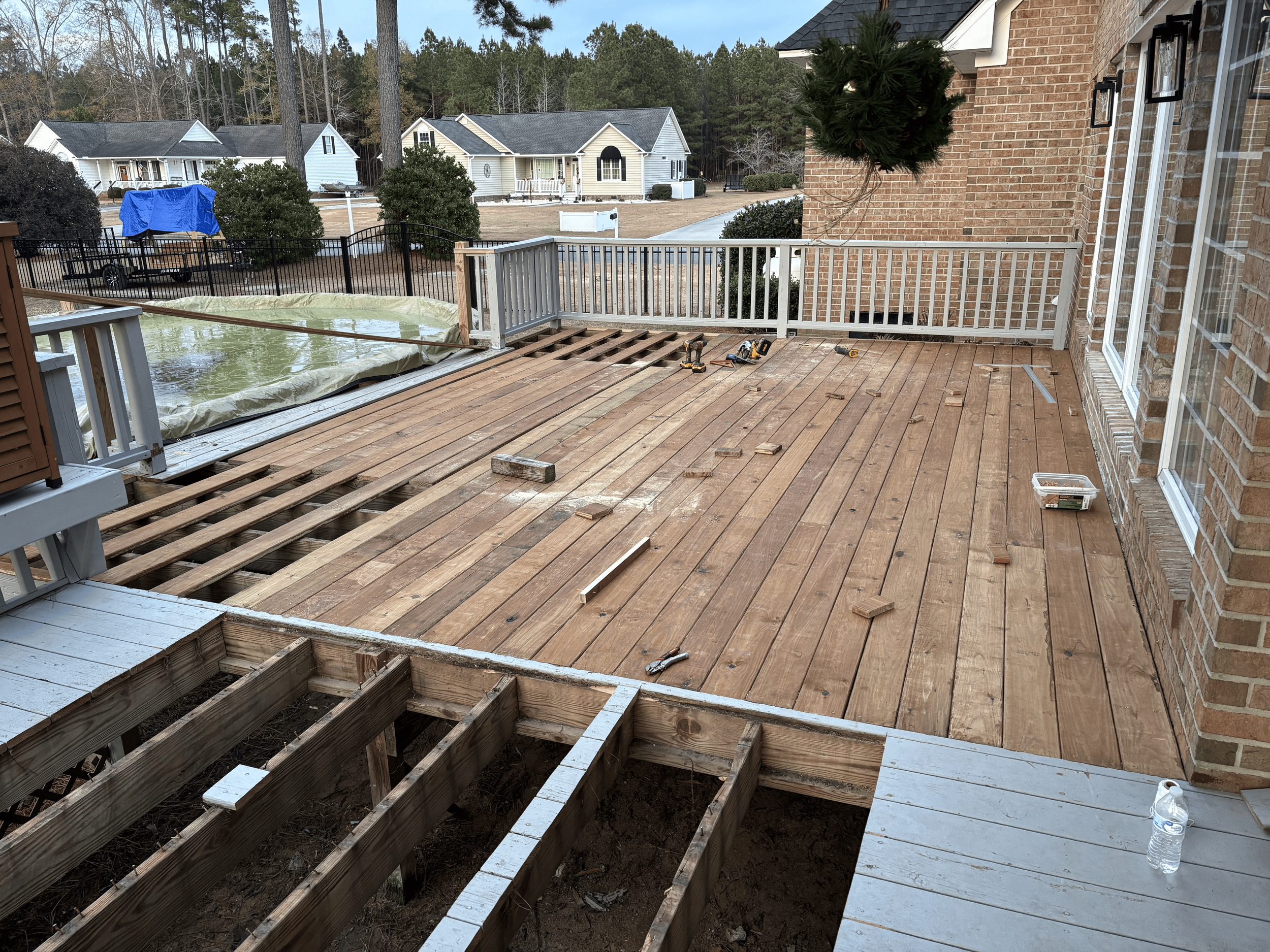 A wooden deck under renovation with some flooring unfinished, tools laying on the deck, and a plastic water bottle on the ground. The backyard has a black fence, trees, and nearby houses, with one house covered in a blue tarp.