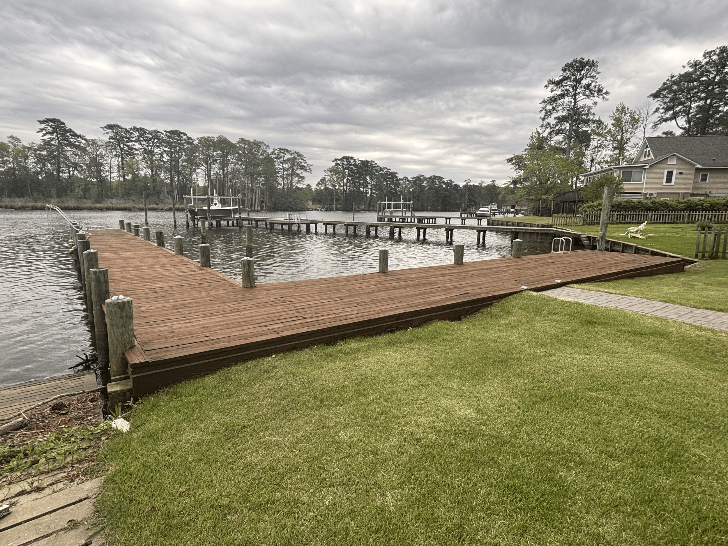 View of a waterfront dock with two boats, grassy lawn, wooden fences, a house, and tall trees under a cloudy sky.