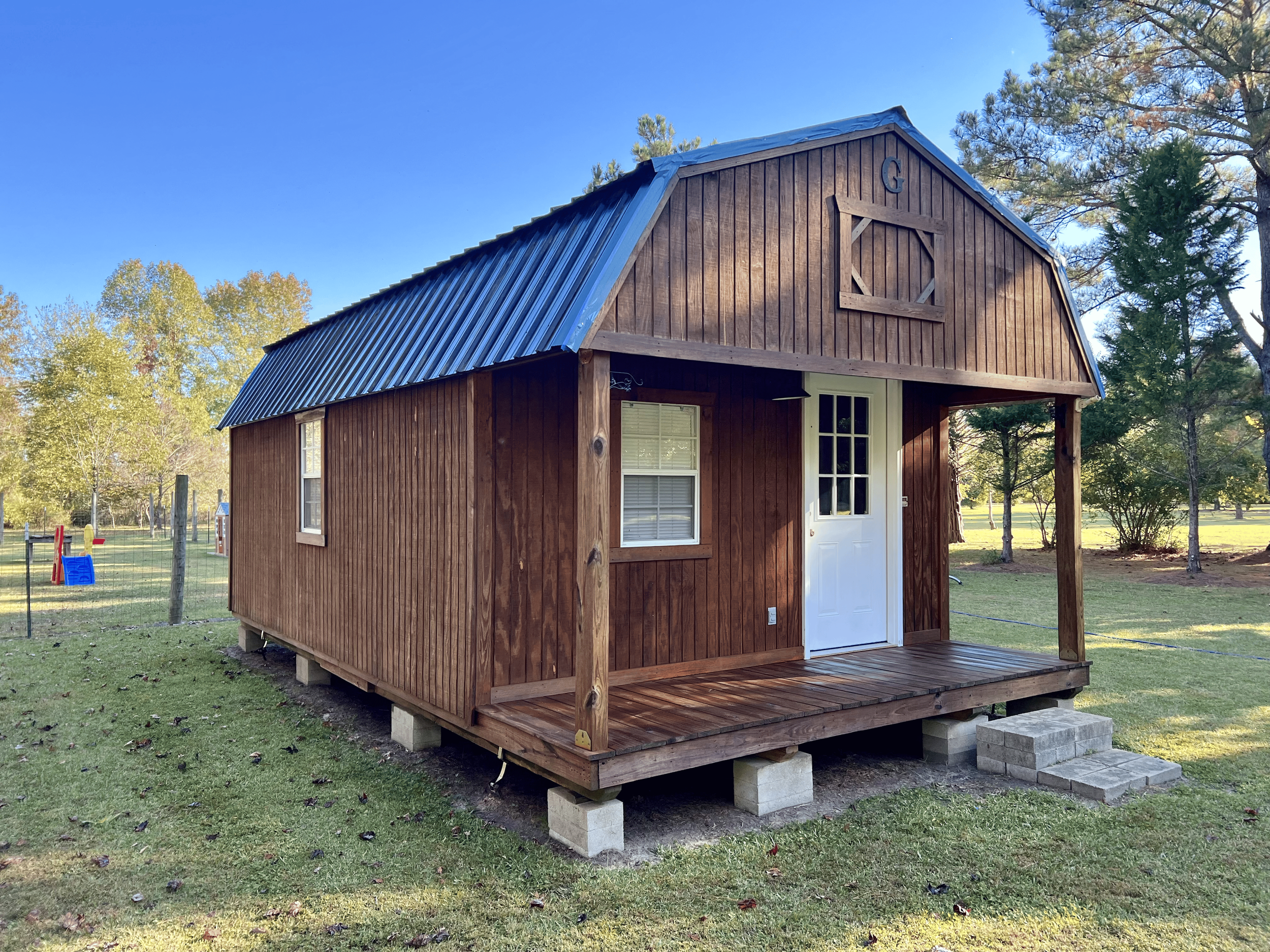 View of a small wooden barn with a steep metal roof, a white door, and windows, set in a grassy area surrounded by trees under a clear blue sky.