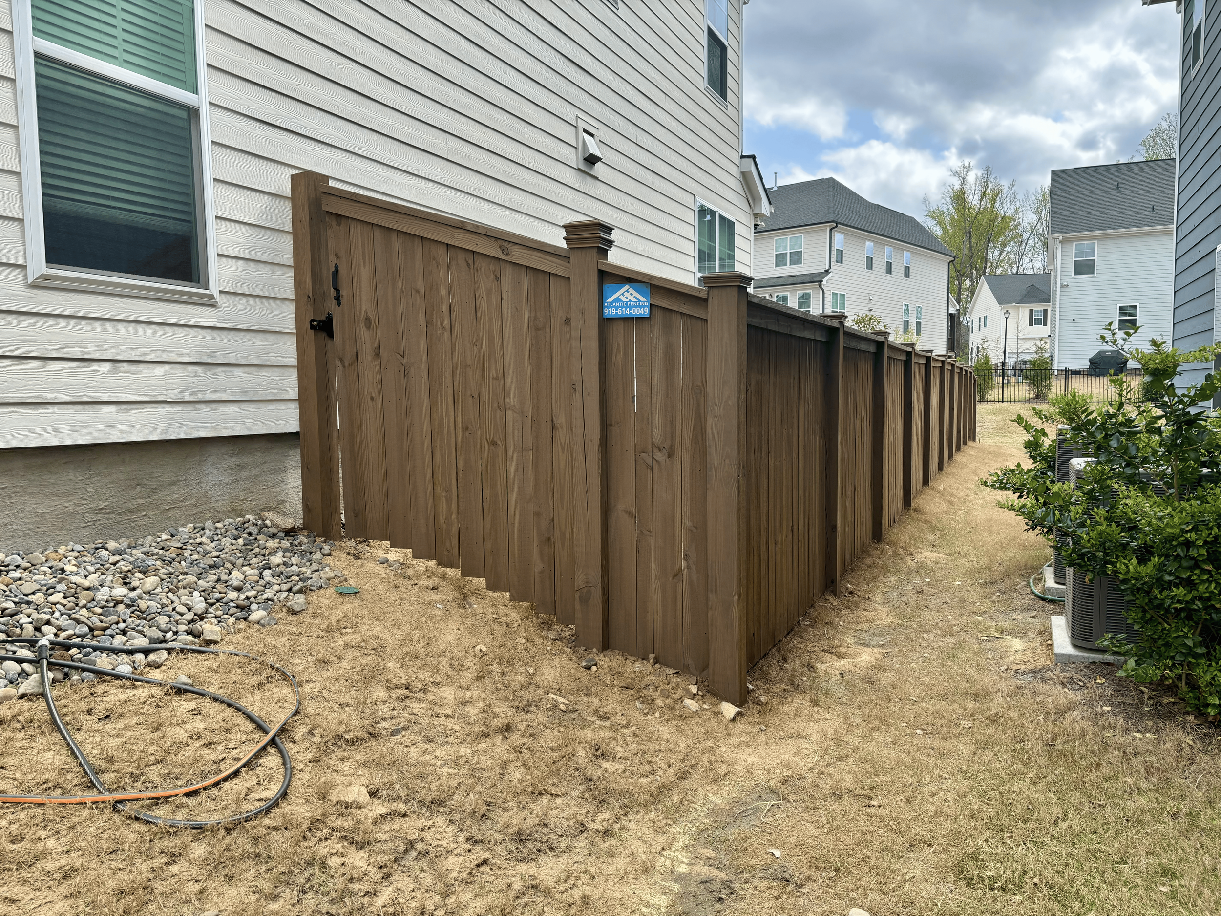 A backyard with a dirt ground and patches of yellow grass, fenced with a wooden privacy fence, with neighboring houses and trees in the background under a cloudy sky.