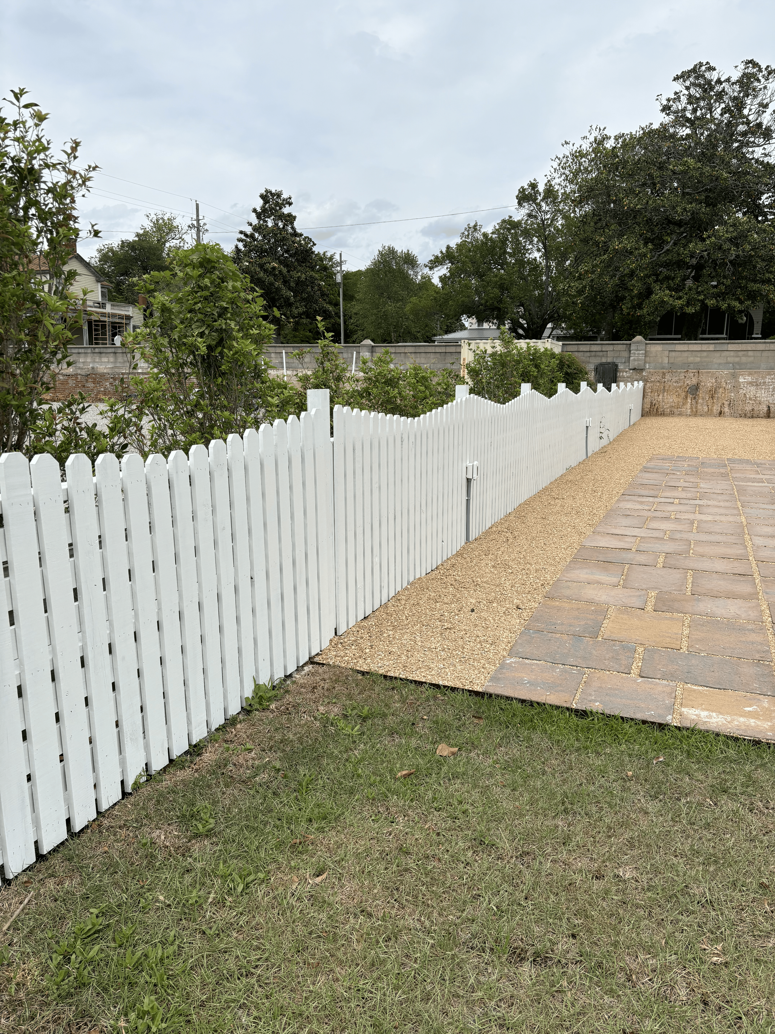 A backyard with a white picket fence, green grass and bushes, a paved walkway, and a concrete wall in the background with trees and houses behind it.