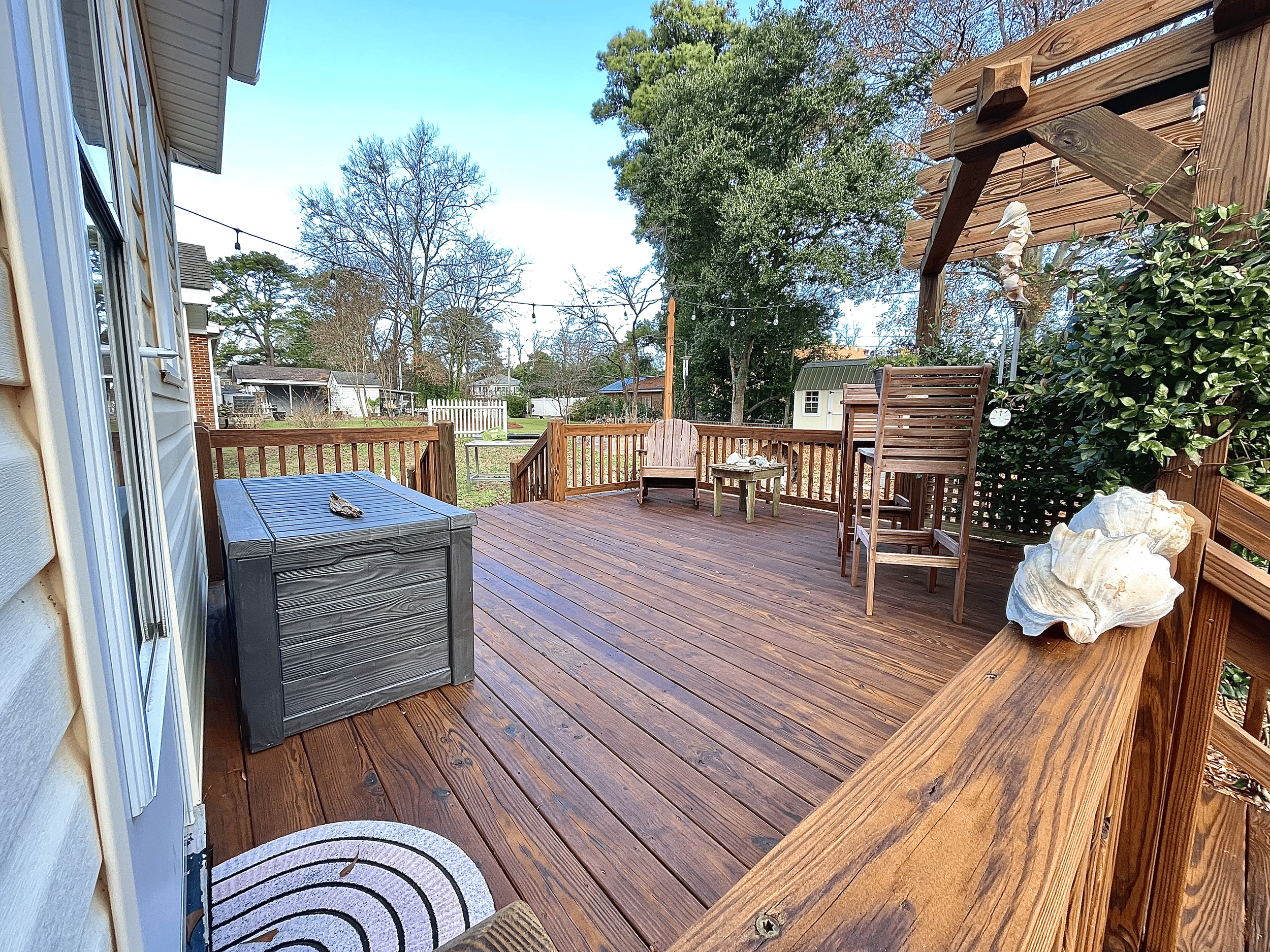 View of a wooden outdoor deck with patio furniture, decorative shells, a small table, and string lights overhead. The background shows trees, a fence, and neighboring houses on a clear day.