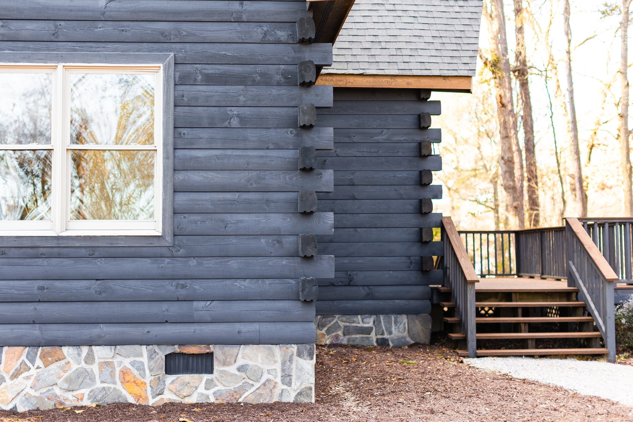 A close-up view of a gray wooden house wall with white-framed windows, stone foundation, and stairs leading to the entrance, with a background of tall trees and sunlight.