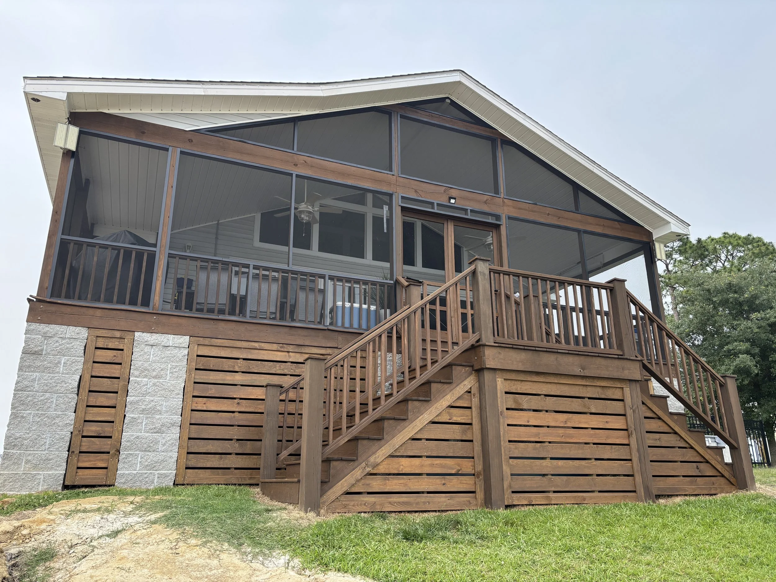 A two-story house with a screened-in porch on the upper level, wooden stairs leading up to it, and a stone foundation. The house has a white roof, and the exterior walls are a combination of stone and wood paneling. There are trees on the right side, and the sky is clear.