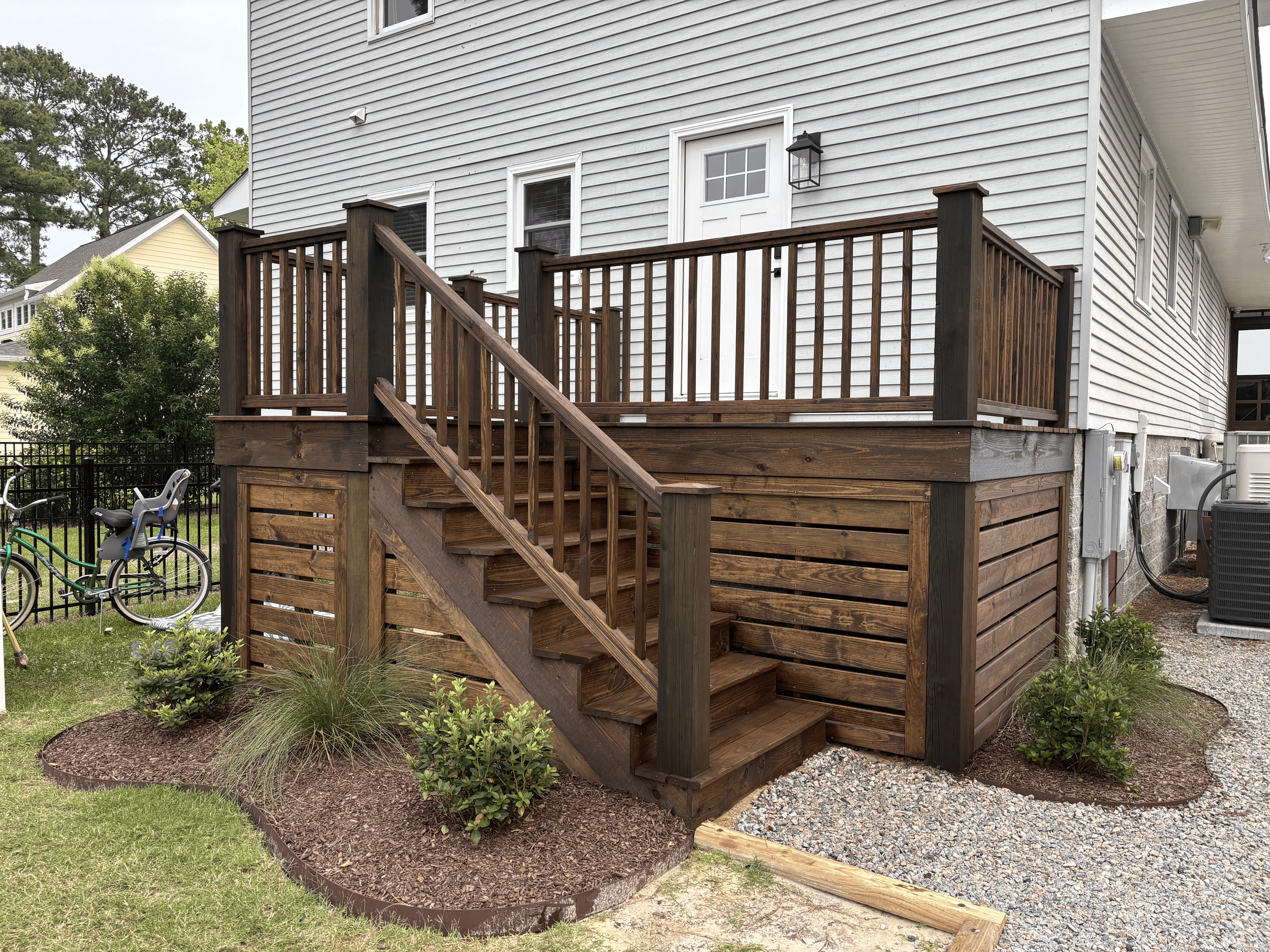 The back porch of a house with wooden stairs, railing, and a white door, surrounded by a landscaped yard with plants and bicycles.