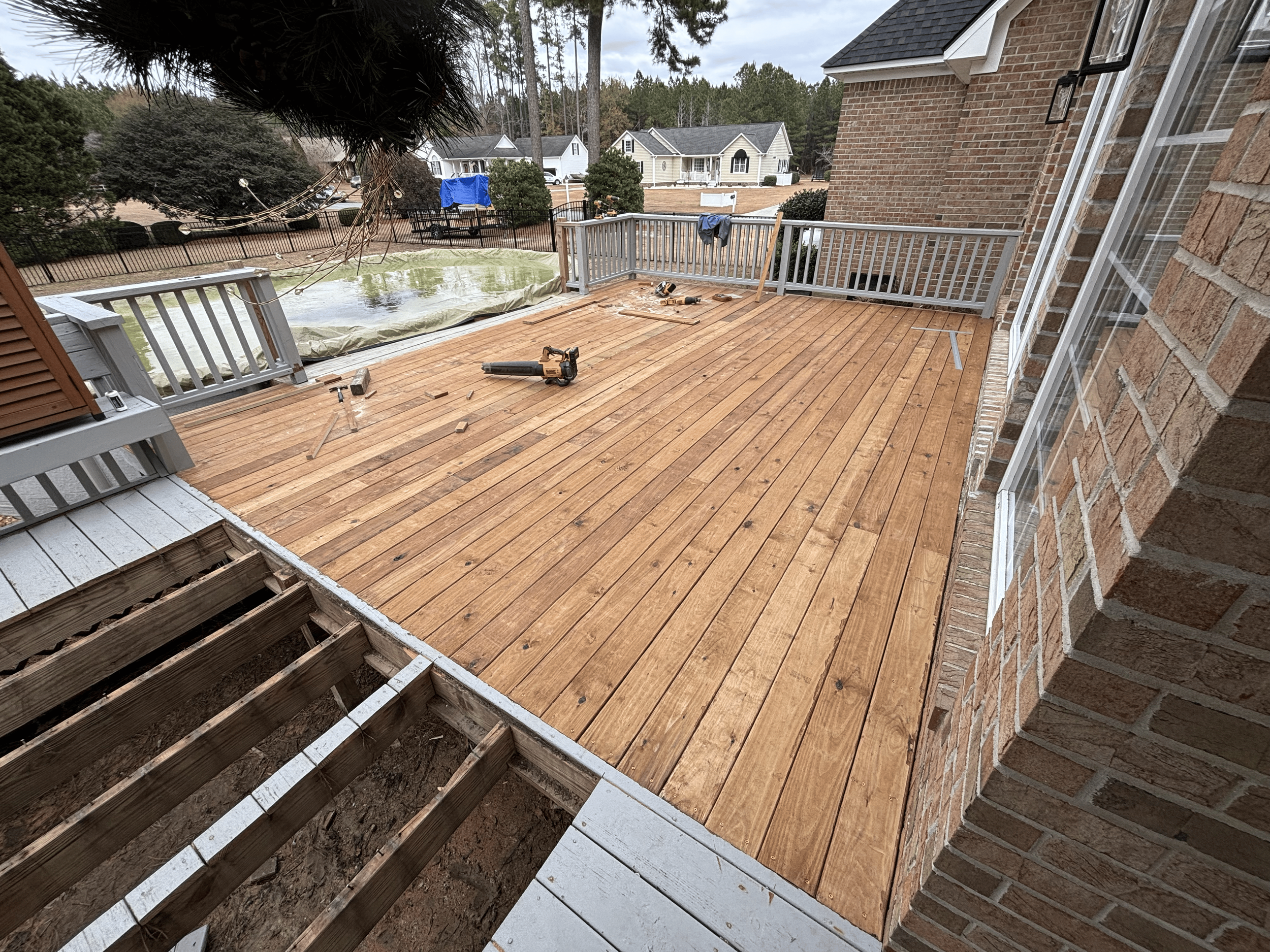 View of a backyard patio under renovation with wooden decking, tools, and construction materials, outside a brick house with neighboring houses in the background.