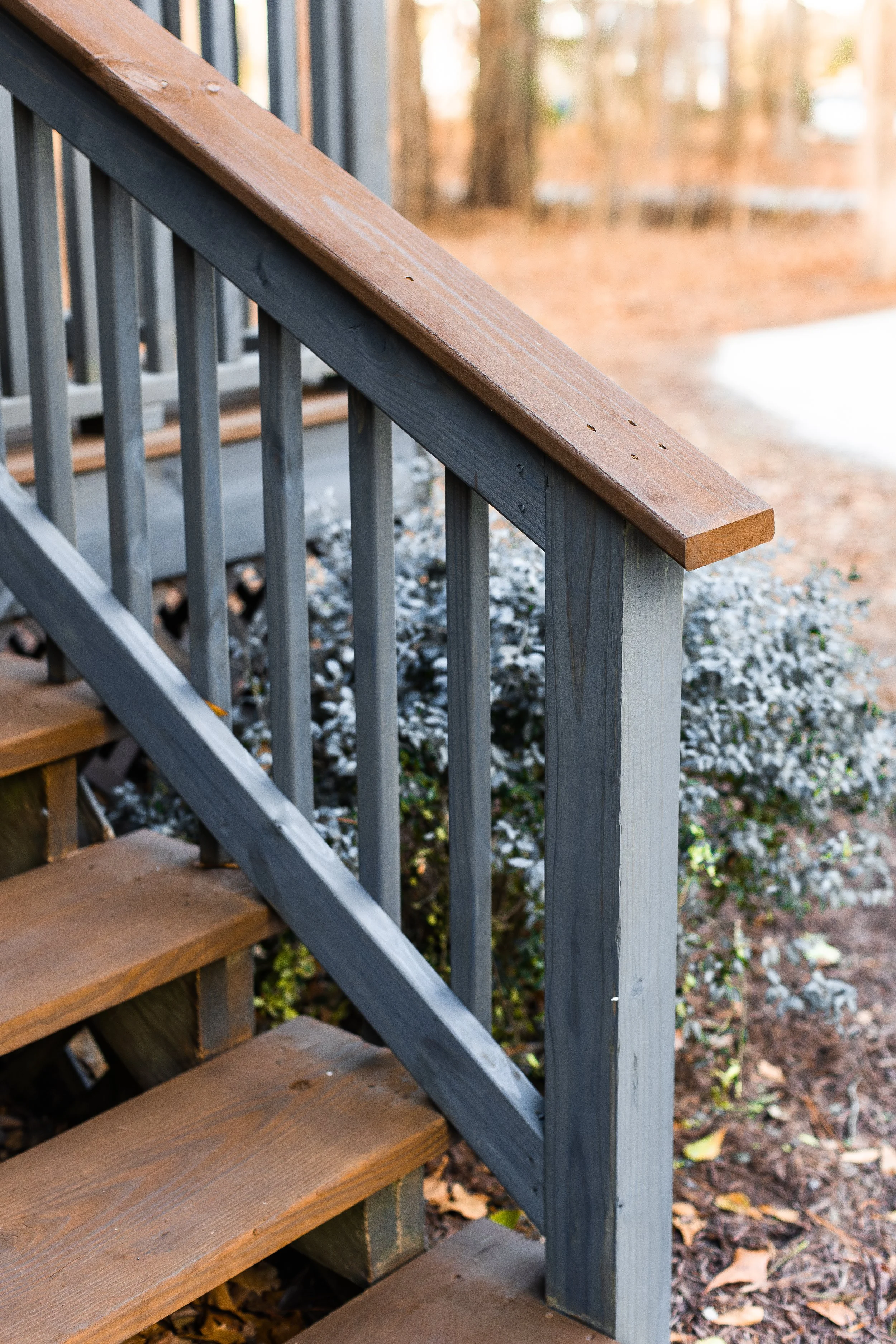Close-up of wooden stairs with gray railing and brown handrail outside, with fallen autumn leaves on the ground.