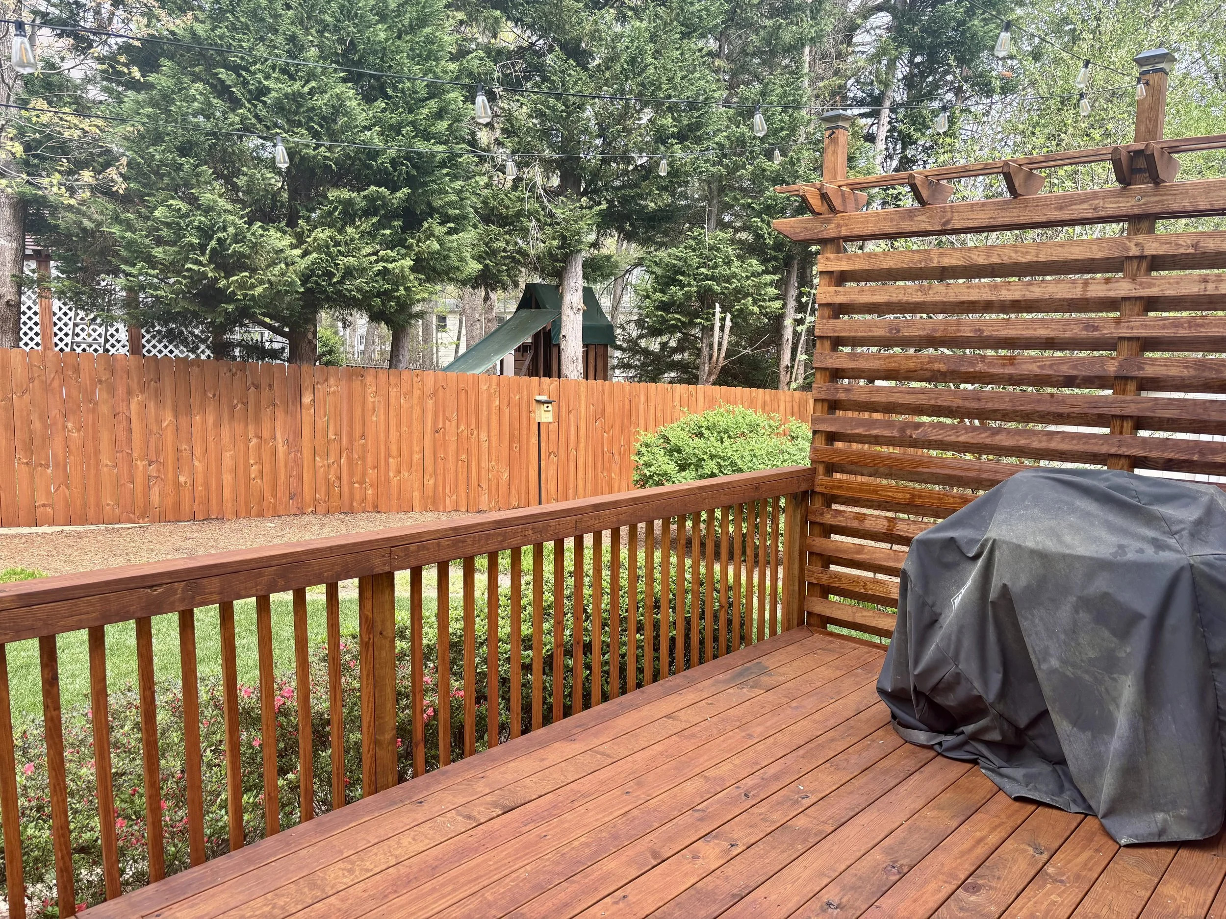 Wooden deck with a black covered grill in the lower right corner, a wooden fence with a small birdhouse, tall trees with green foliage, a playground slide in the background, and string lights hanging overhead.