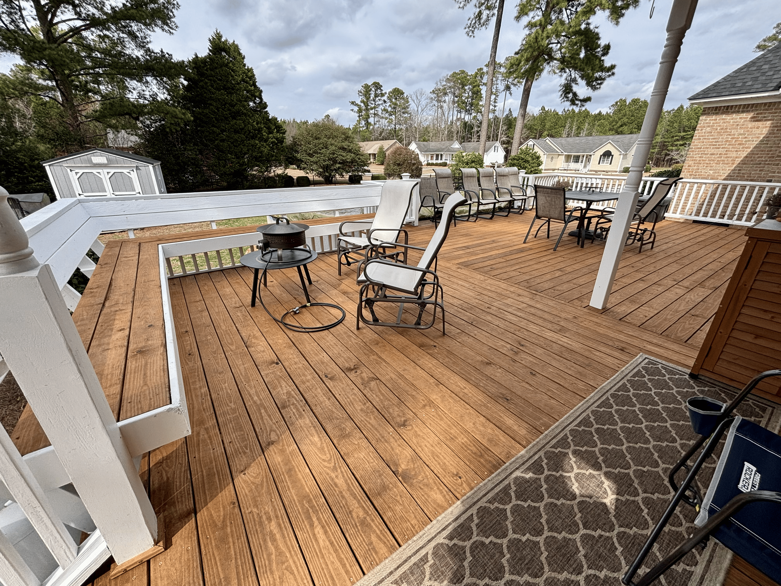 View of a backyard wooden deck with patio furniture, including several chairs and a table, surrounded by trees and neighboring houses under a cloudy sky.