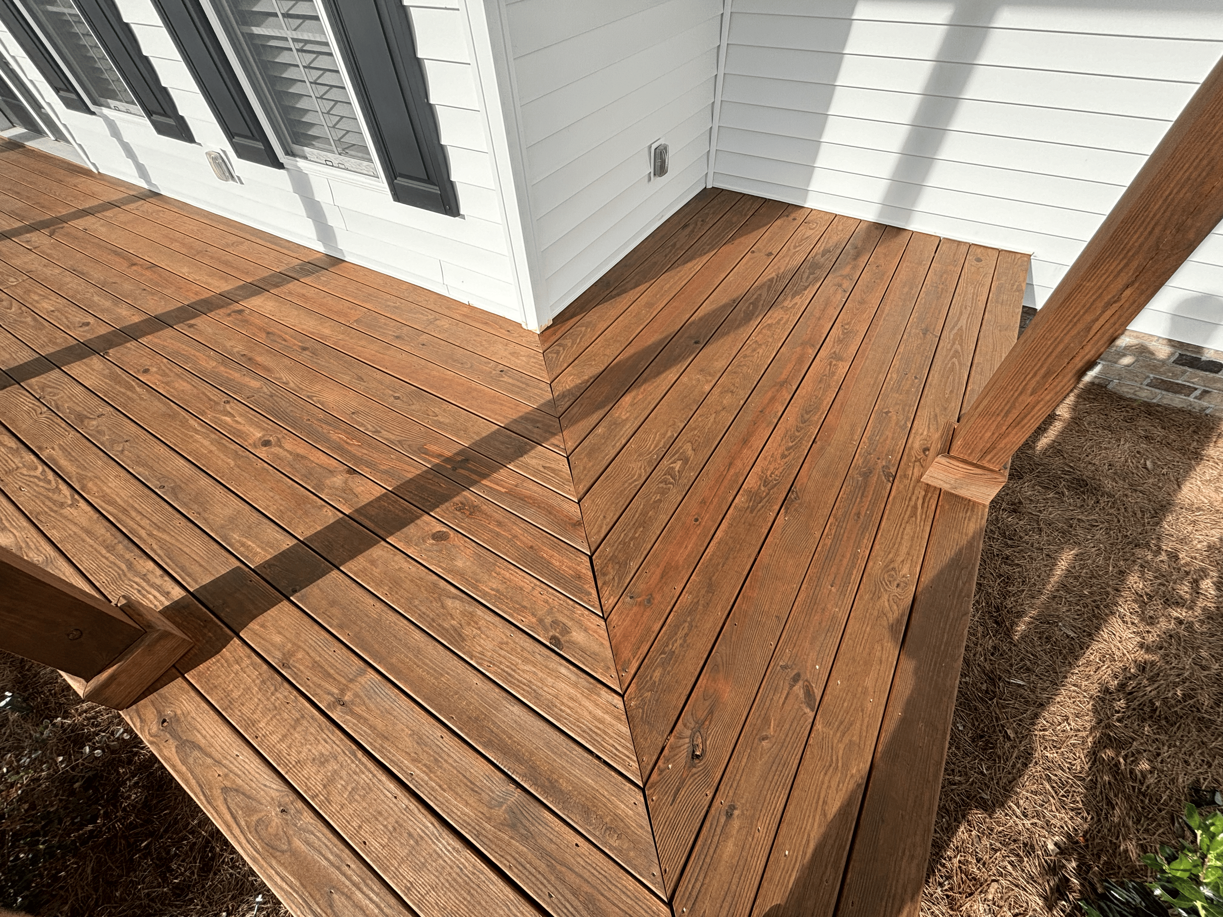 A newly built wooden deck attached to the side of a white house with black window shutters, shadows cast by sunlight, and some dirt and grass seen beside the deck.