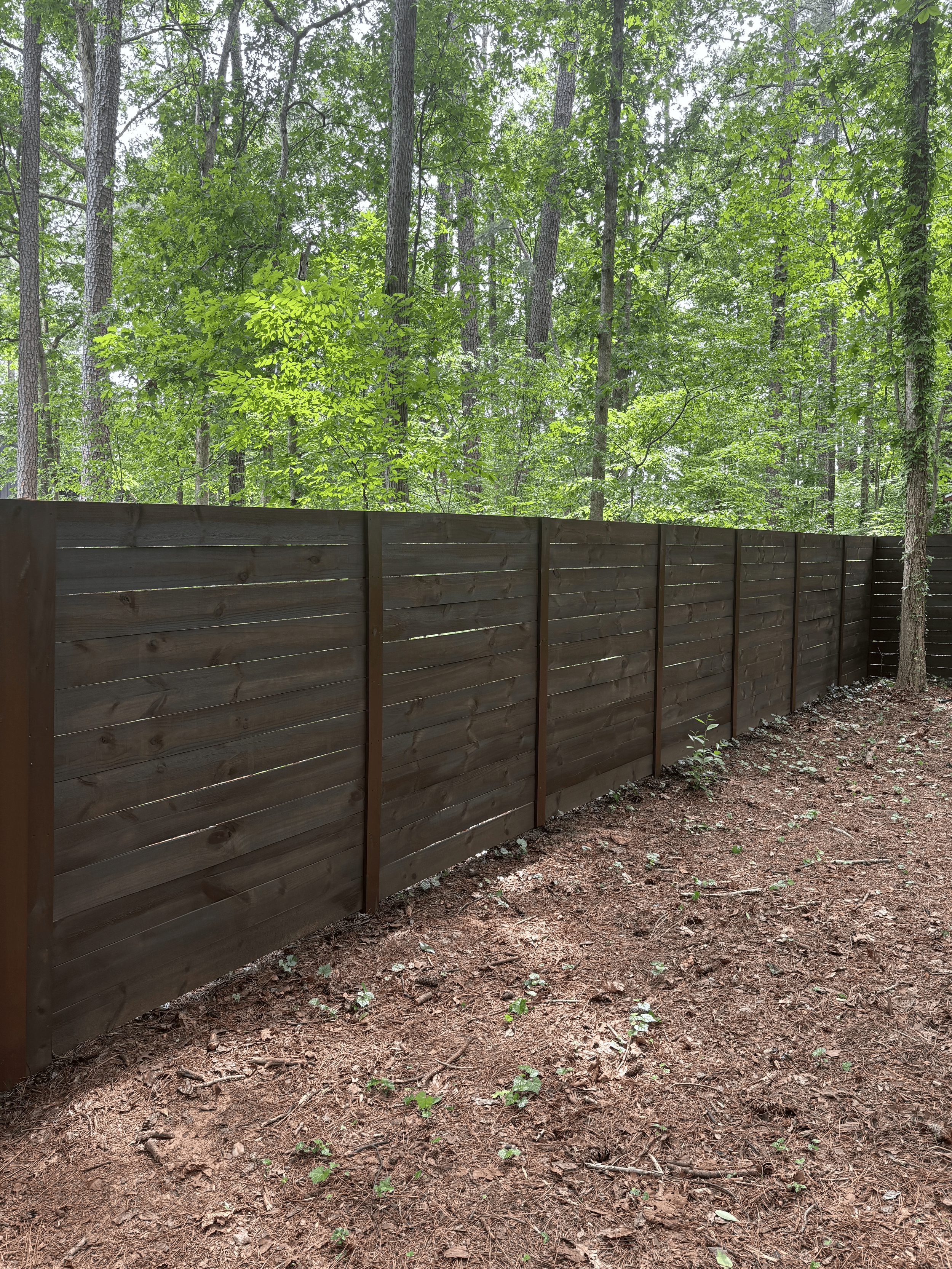 Wooden privacy fence in front of a green forest with tall trees and leaves, on a dirt ground