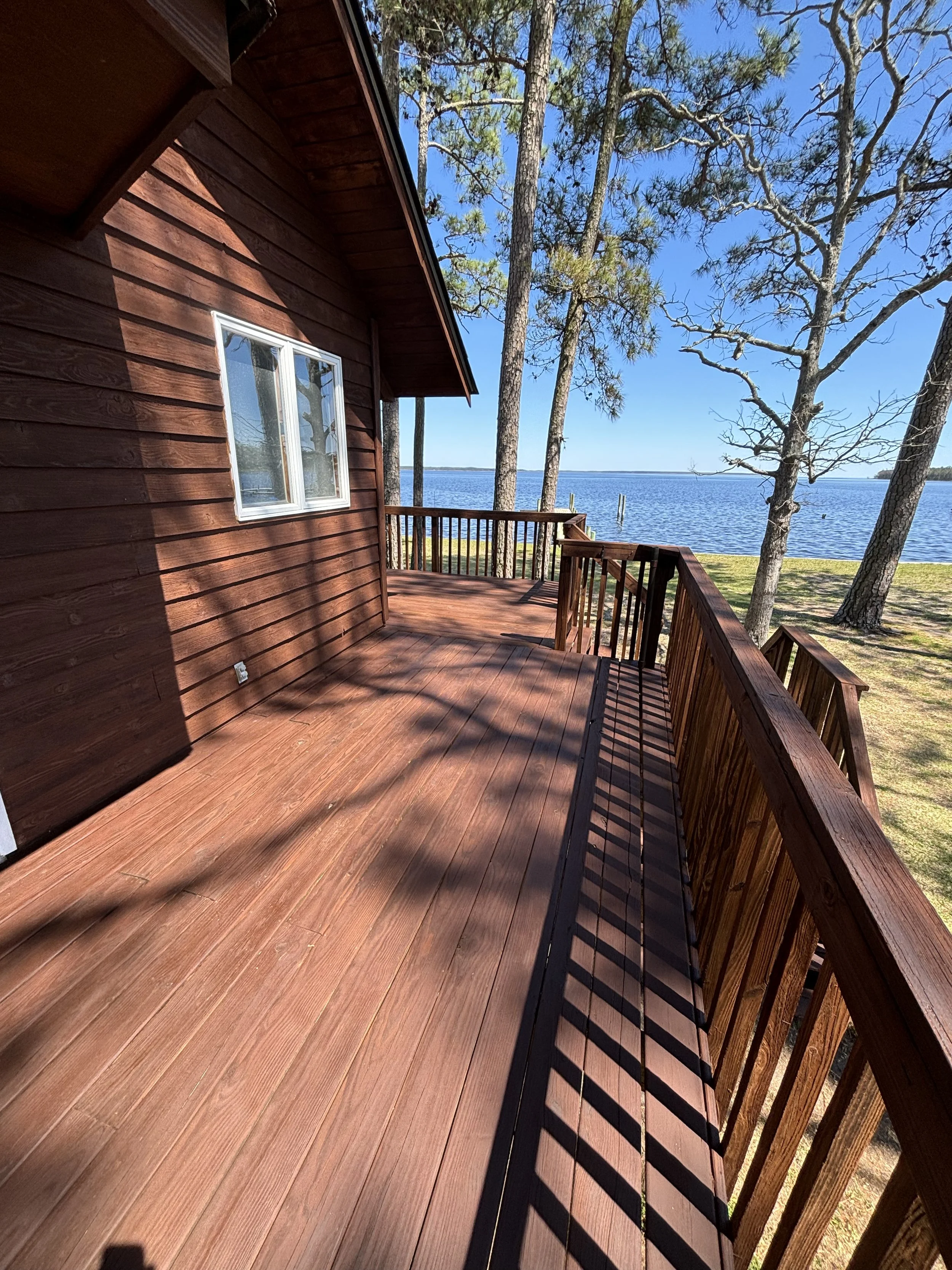 A wooden deck with a railing overlooking a lake, with tall trees and a clear blue sky in the background.