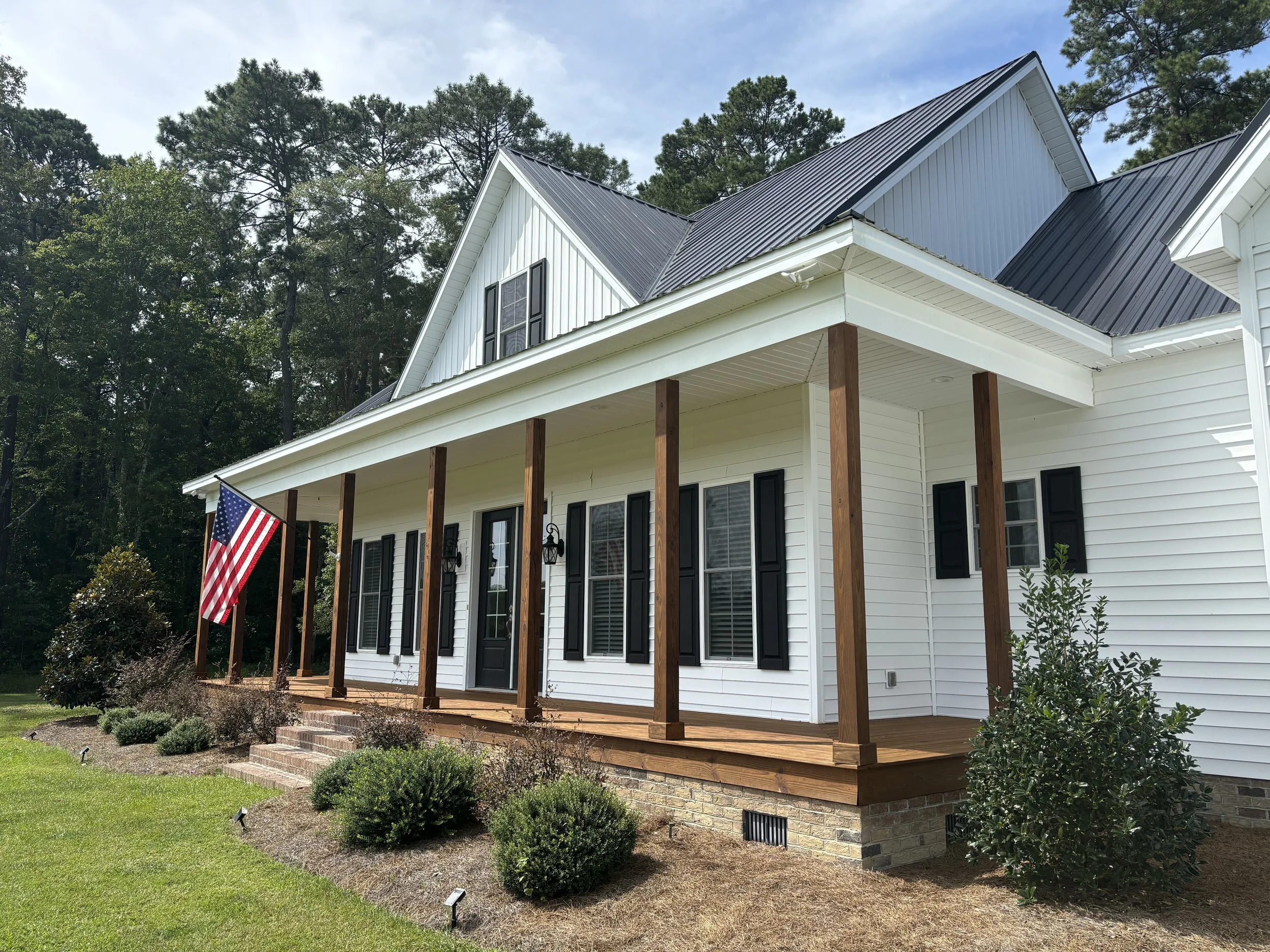 Front view of a white house with black shutters and a black metal roof, featuring a porch with wooden posts and a small set of brick steps, surrounded by lush greenery and an American flag.