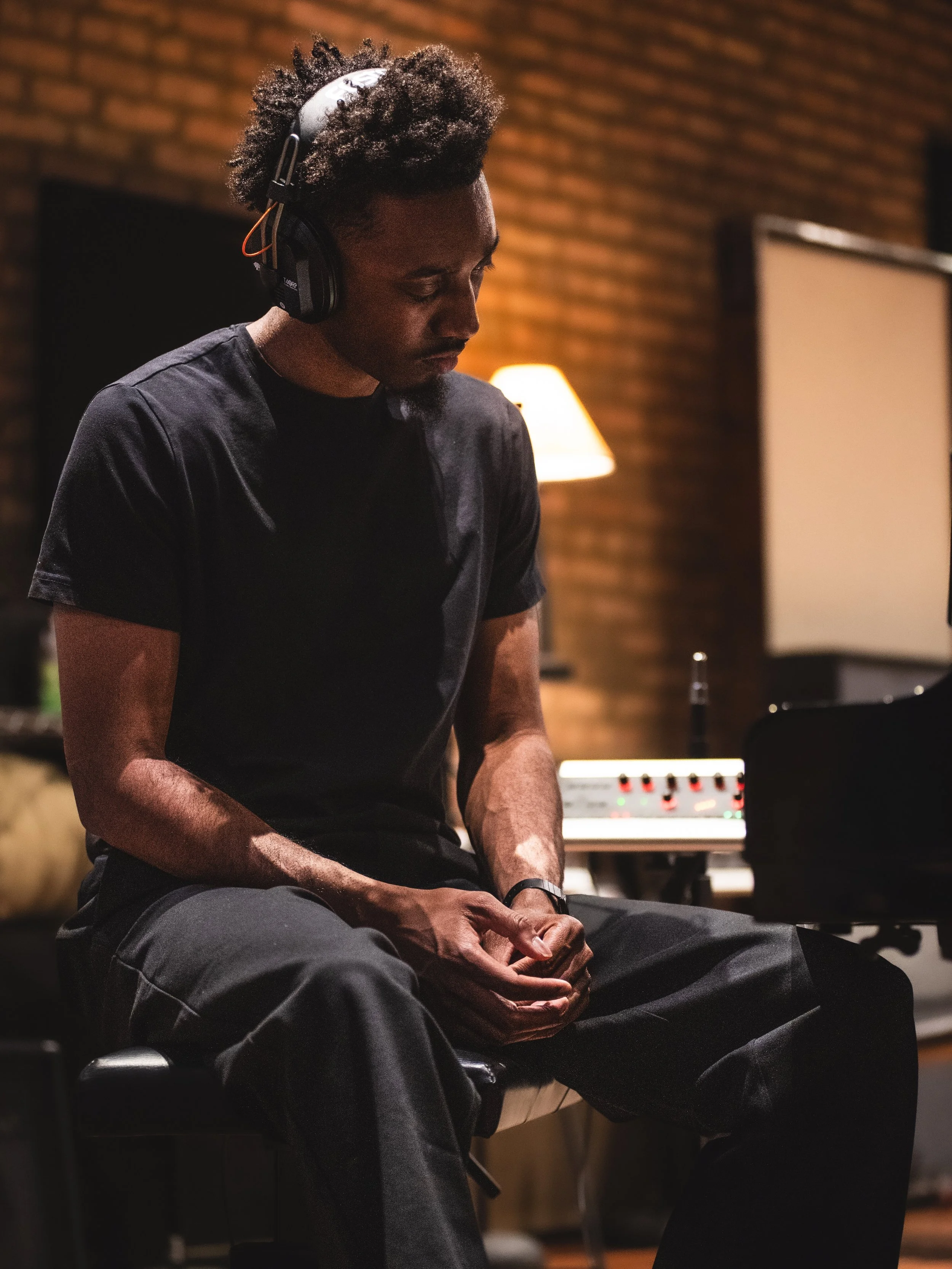 A man sitting in a music studio with headphones, wearing a black T-shirt and pants, and sitting with his hands clasped, in front of music equipment and a brick wall background.