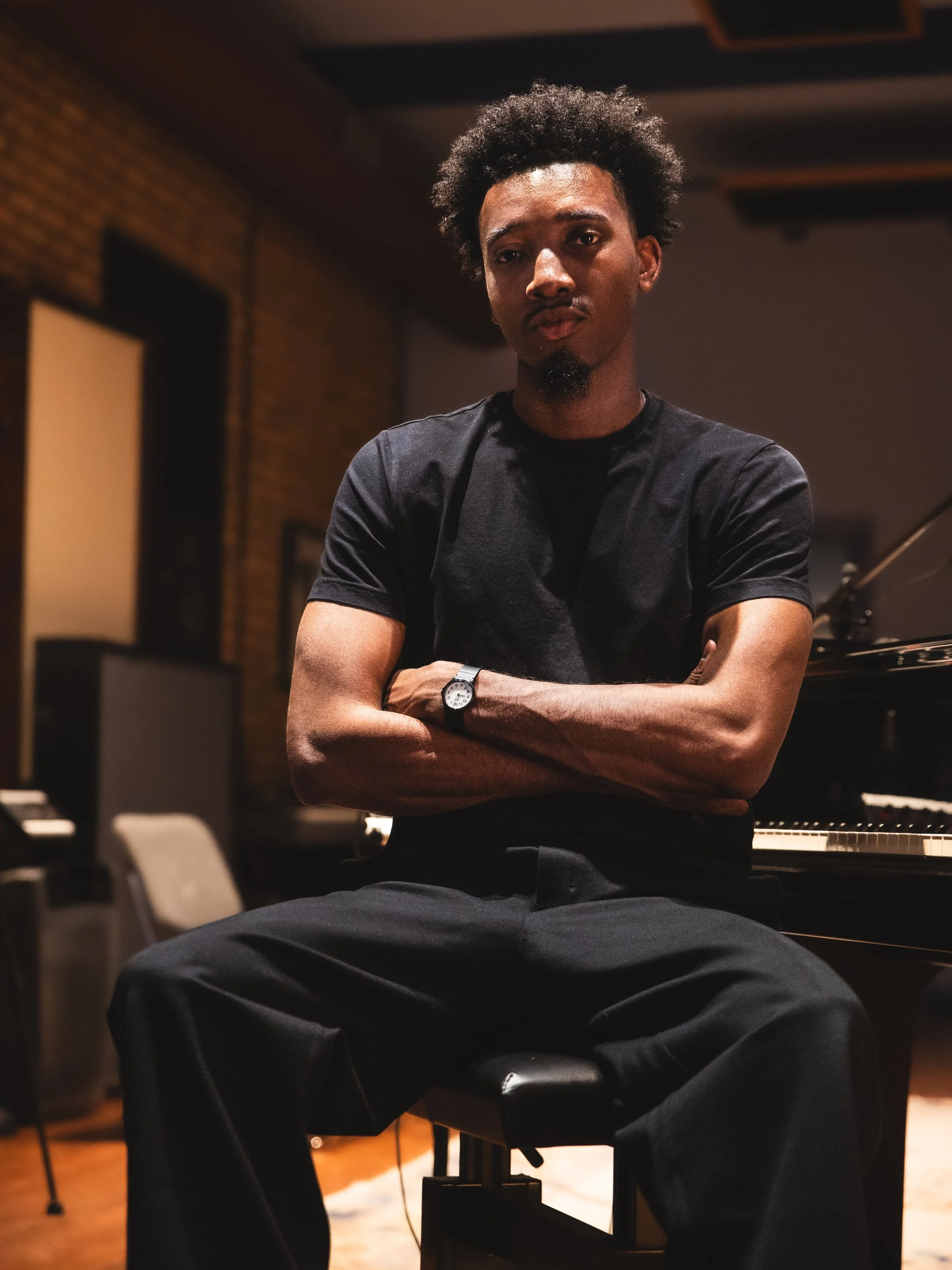 A man sitting on a piano bench with arms crossed, in a music studio or rehearsal space, wearing a black t-shirt and black pants.