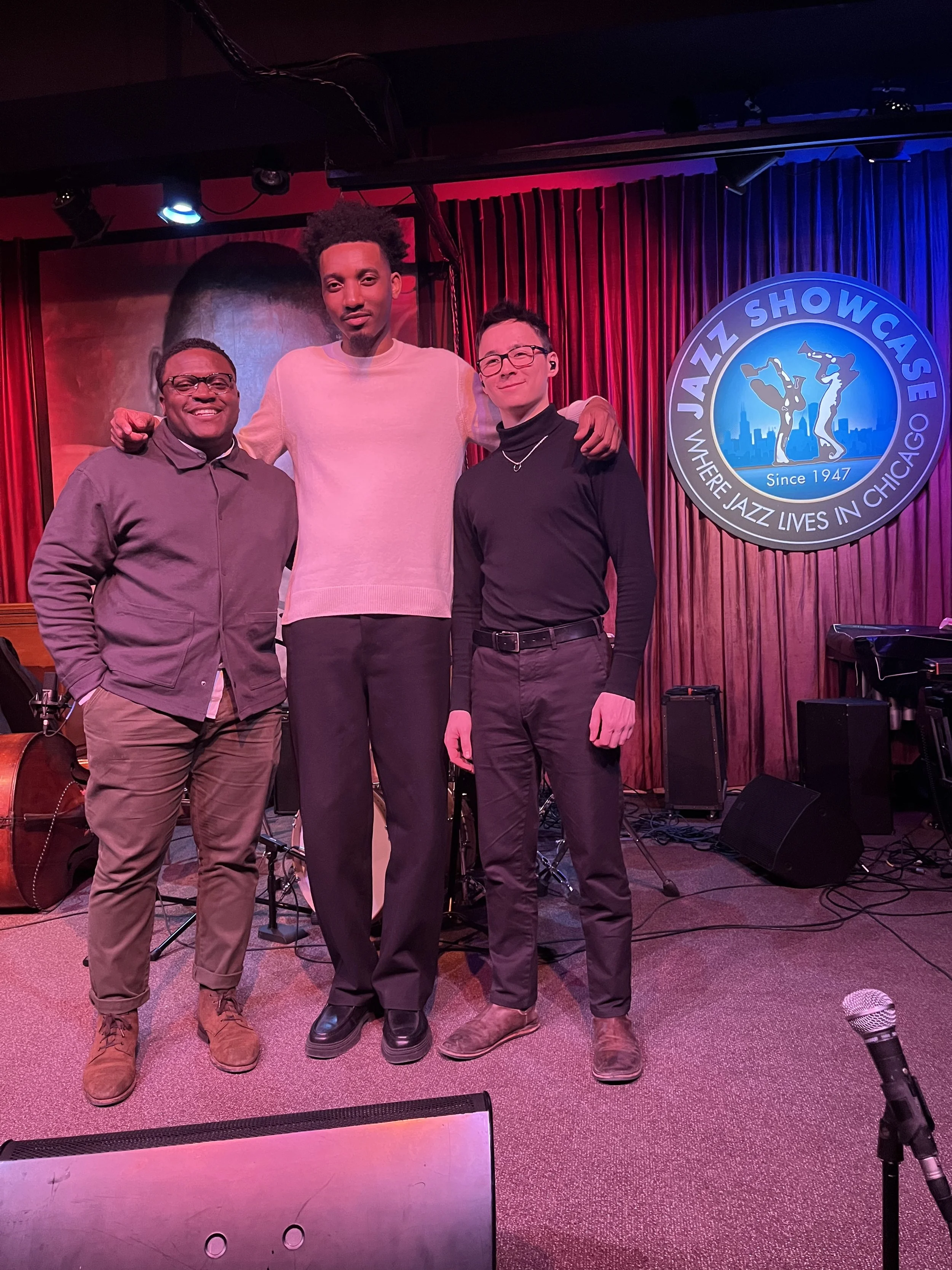 Isaiah Jones Jr. , Sid Smith IV, and Nat Lin posing on stage for a photo at Jazz Showcase in Chicago, with musical instruments and equipment behind them, and a red curtain.