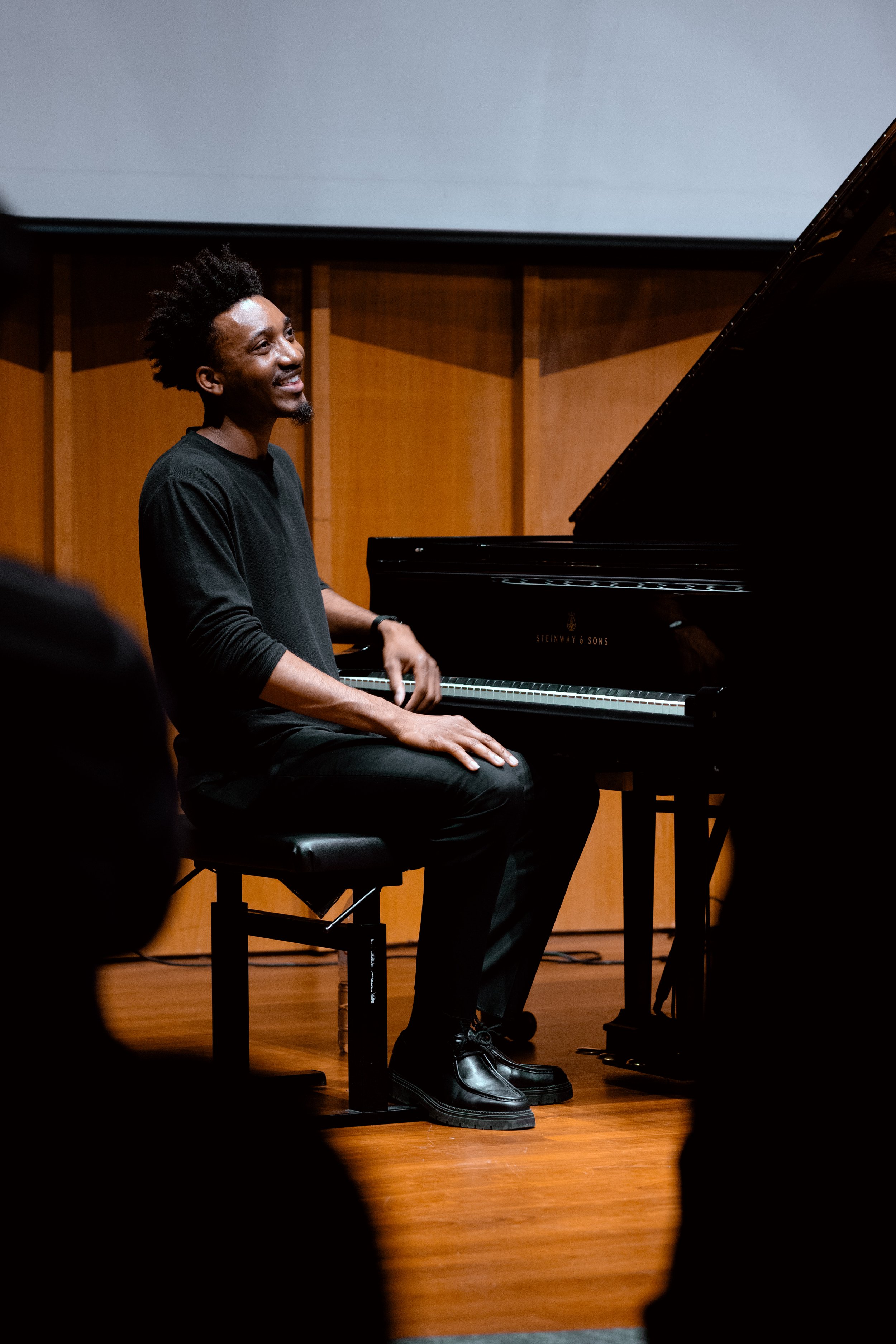 A man sitting at a grand piano on stage, smiling, in a black shirt and pants, with a wooden background behind him.