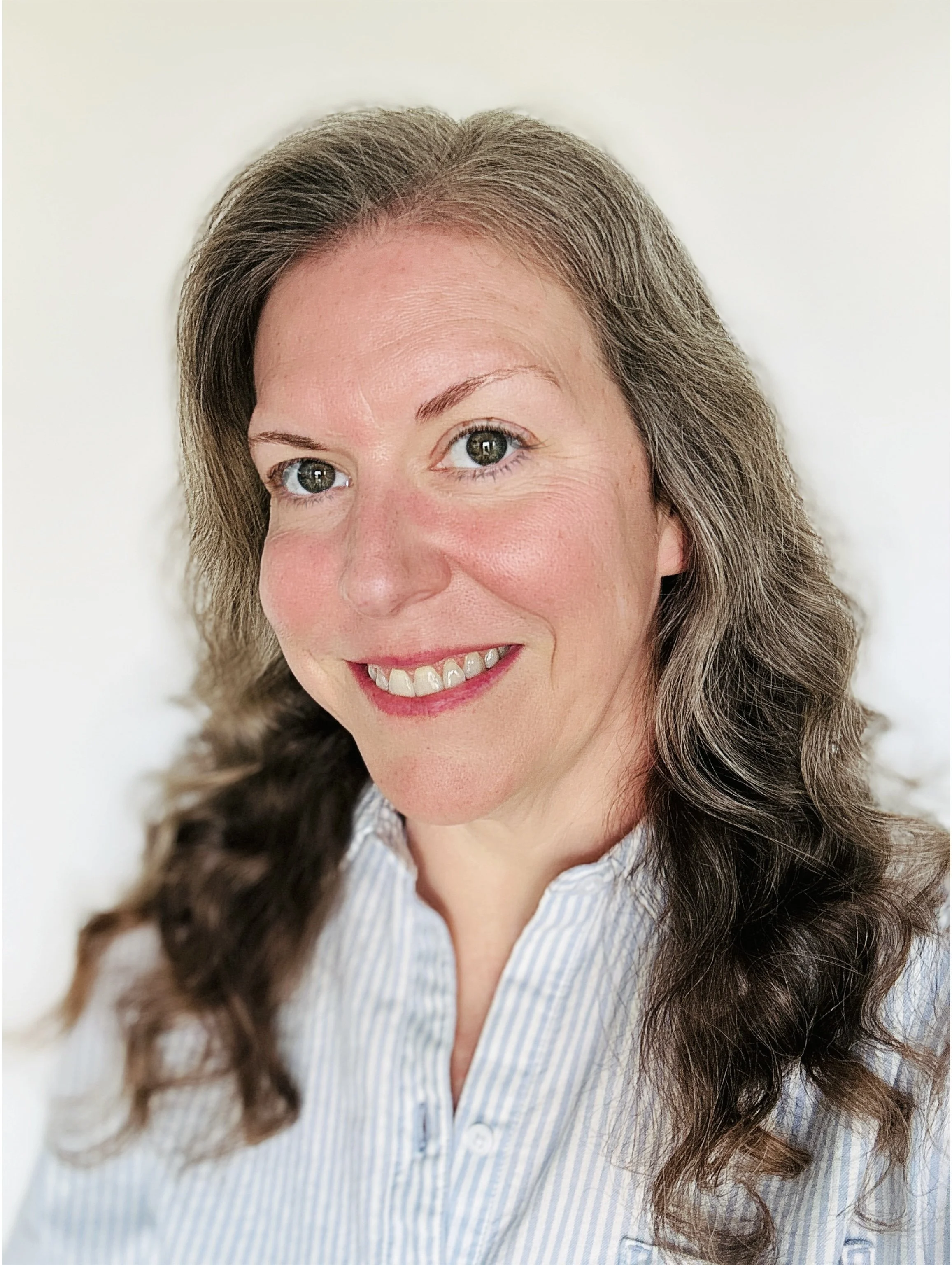 A woman with light skin and curly brown hair smiling, wearing a light blue and white striped shirt, standing against a plain white background.