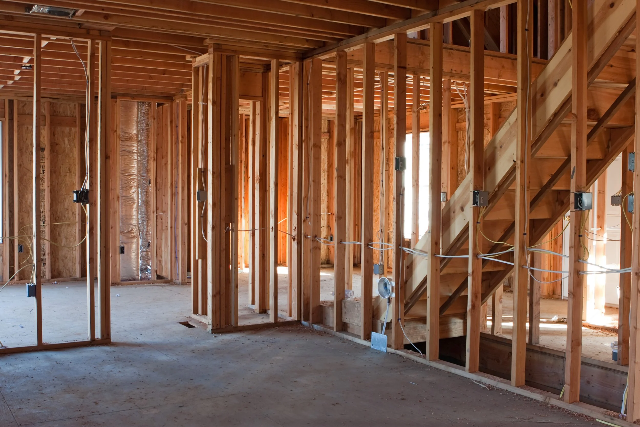 The image shows the interior of a house under construction, with exposed wooden framing, electrical wiring, and staircases.