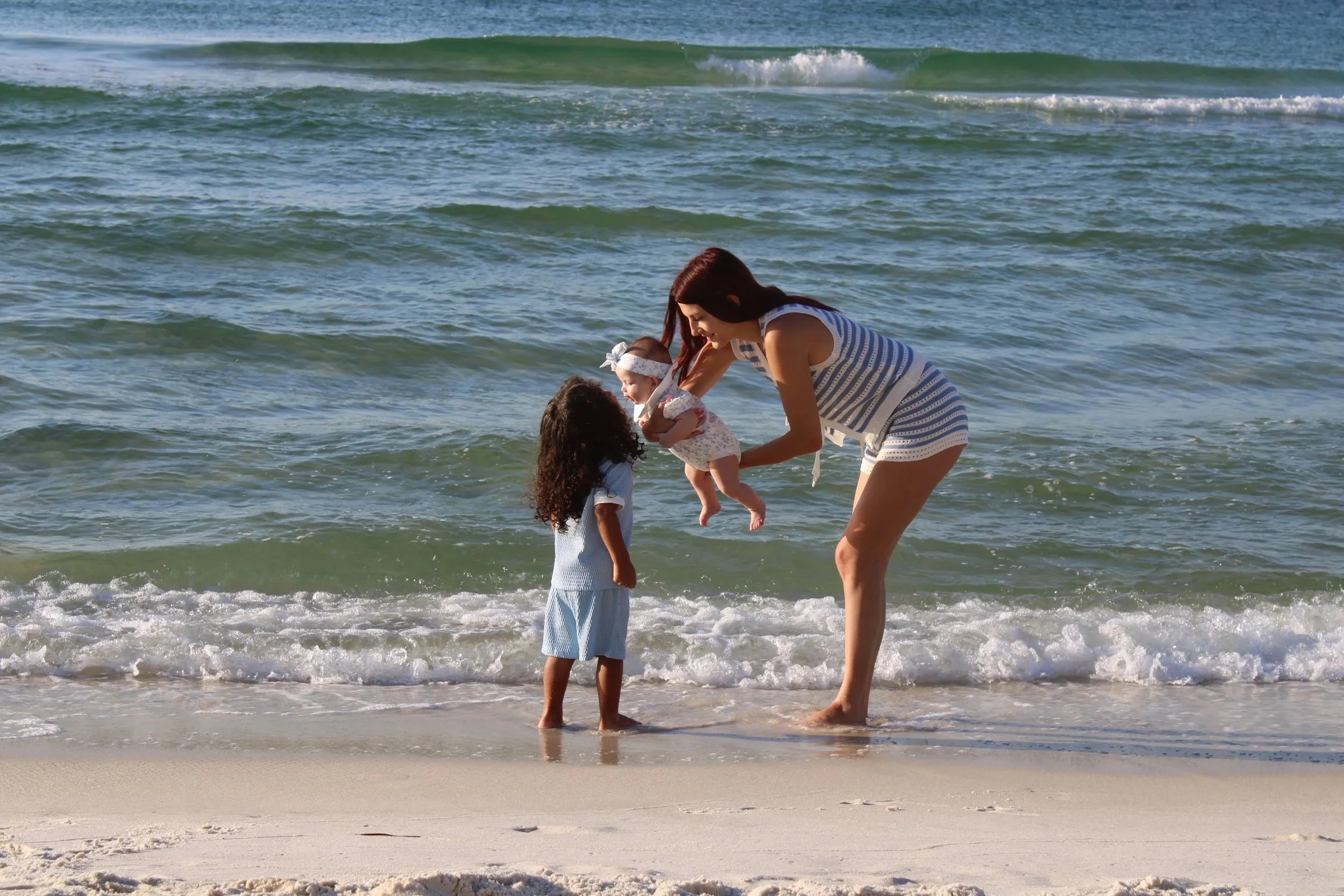 A woman and two young girls at the beach, with the woman holding a baby girl, as they stand near the shoreline with waves in the background.