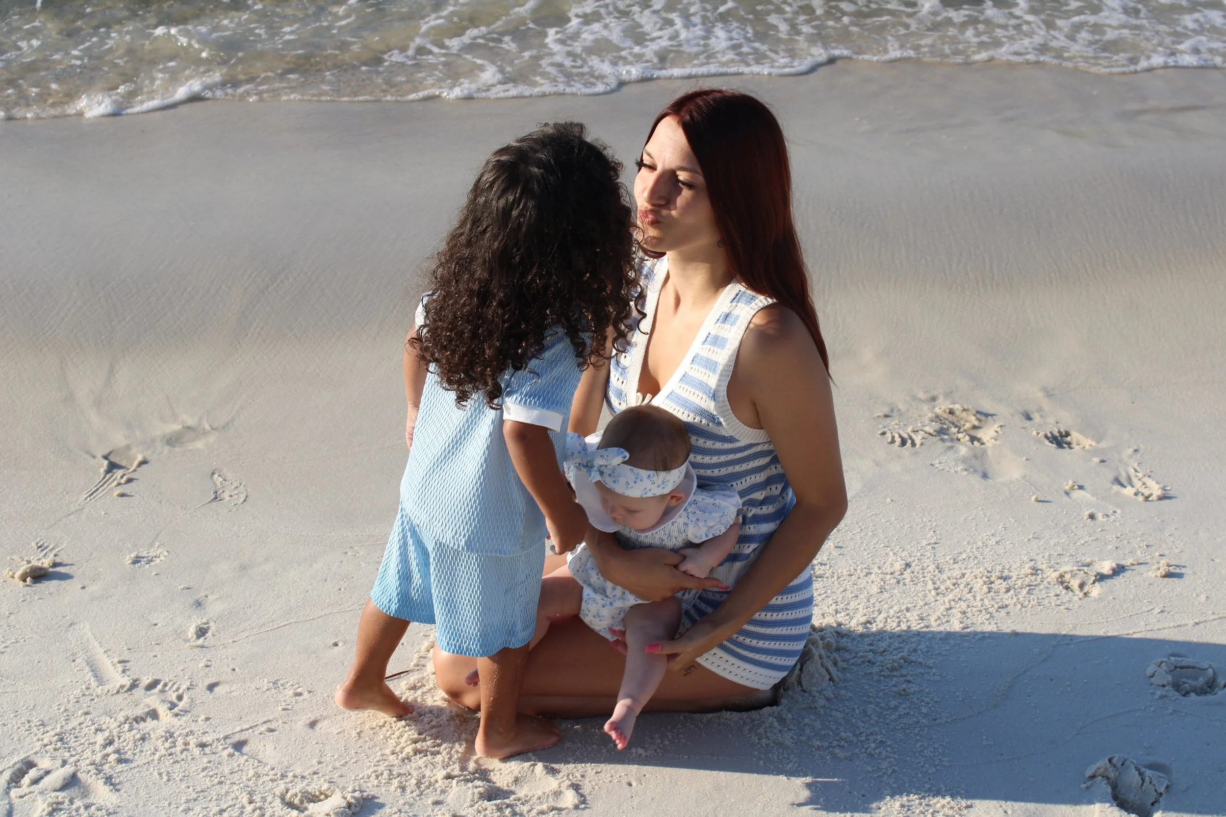 A woman with red hair kneeling on the sand at the beach holding a baby dressed in white. A young girl with curly hair is kissing the woman on the lips. The ocean waves and footprints in the sand are visible in the background.