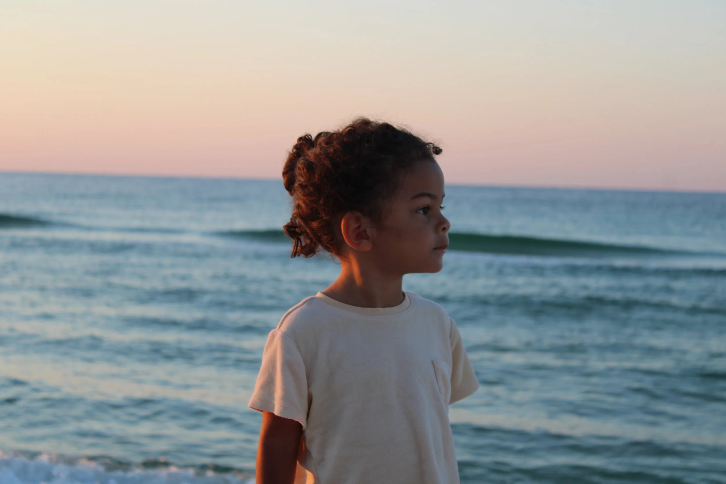 A young boy with curly hair standing on the beach, looking at the ocean during sunset.