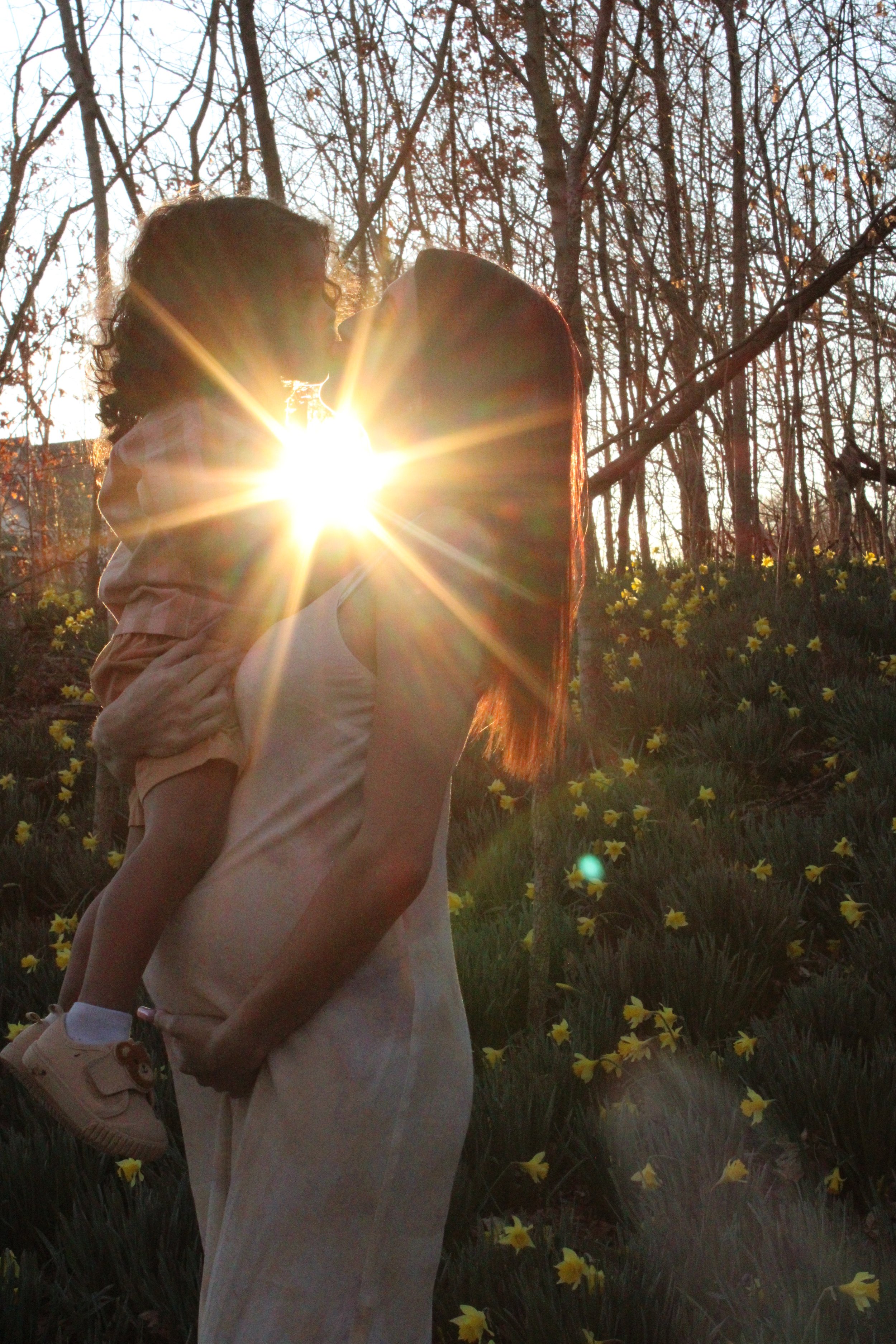 A woman holding a child outdoors during sunset, with trees and yellow flowers in the background.