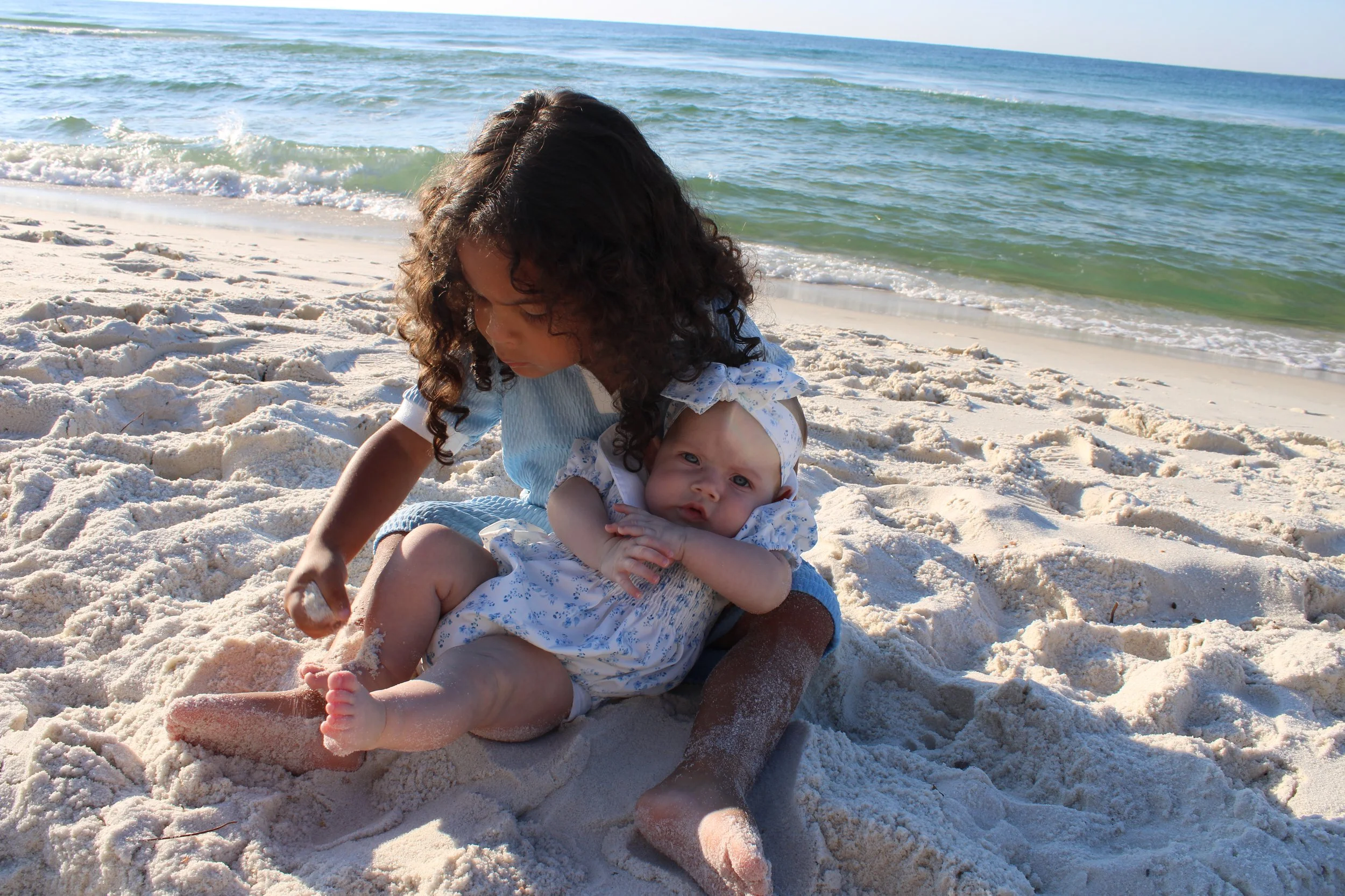 Two young children, a girl with curly hair and a younger girl with a bow in her hair, sitting on sandy beach near ocean with waves in the background.