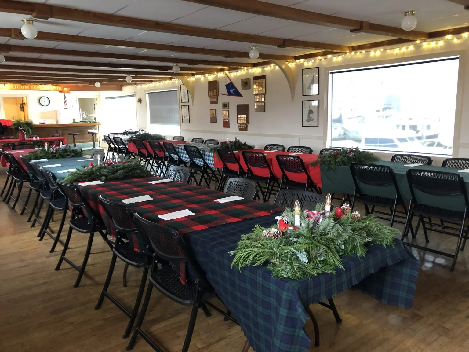 A decorated indoor dining area with long tables covered in holiday-themed tablecloths, pine branch centerpieces, and arranged chairs. The room has wooden beams on the ceiling, string lights, and large windows with a view of boats outside, indicating it's near a marina or harbor.