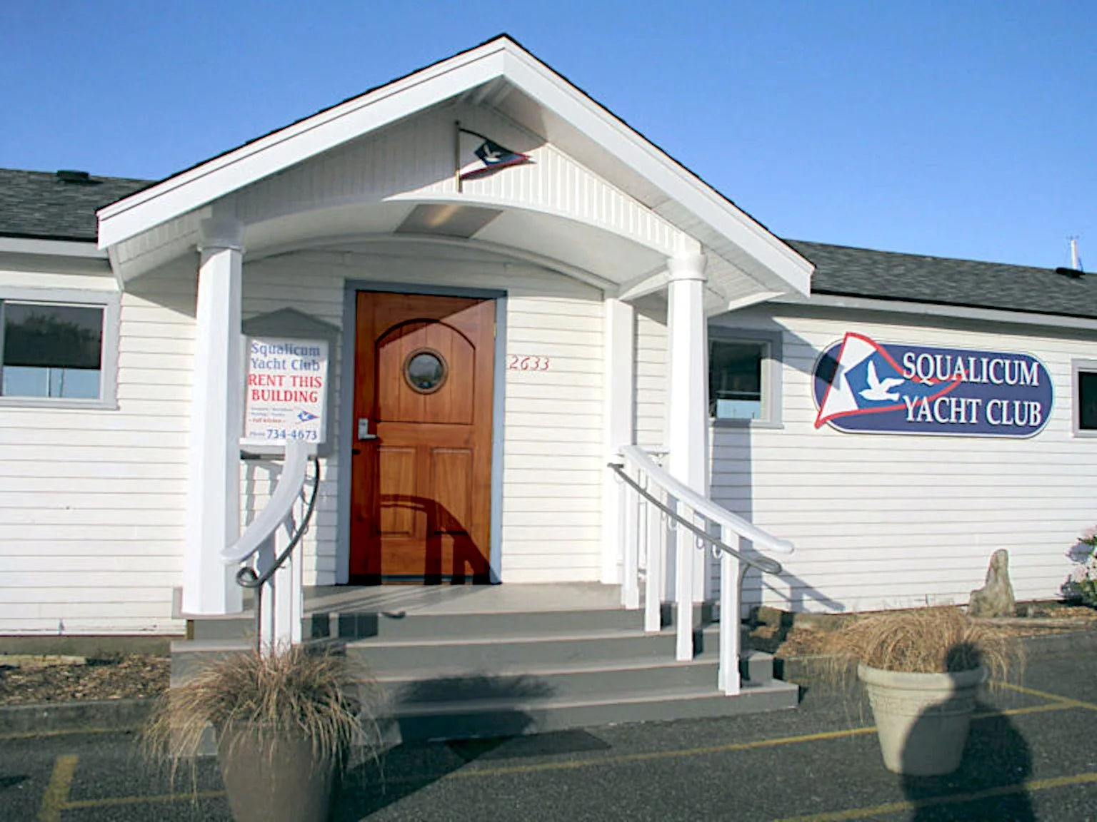 Front of a white building with a wooden door, ramp, and a sign reading 'Squalicum Yacht Club'; also has a sign that says 'Rent this building' with a phone number.