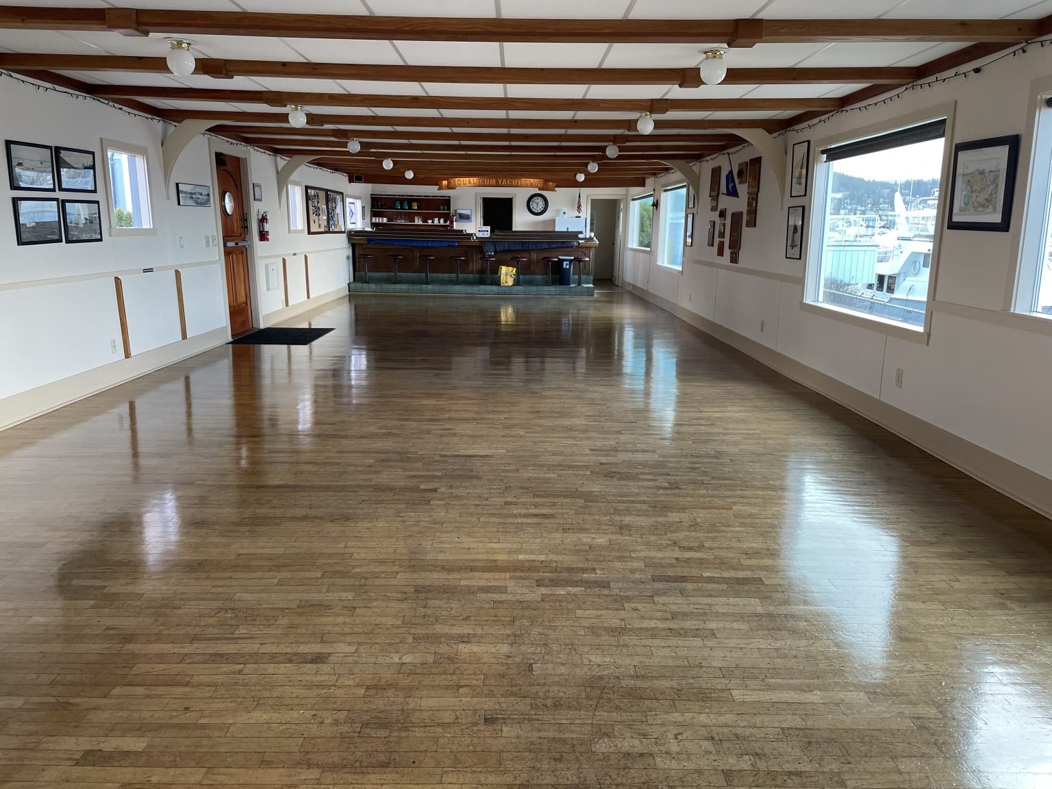 Empty event space with polished wooden floors, white walls decorated with framed pictures, and a bar at the far end, with windows showing boats docked outside.