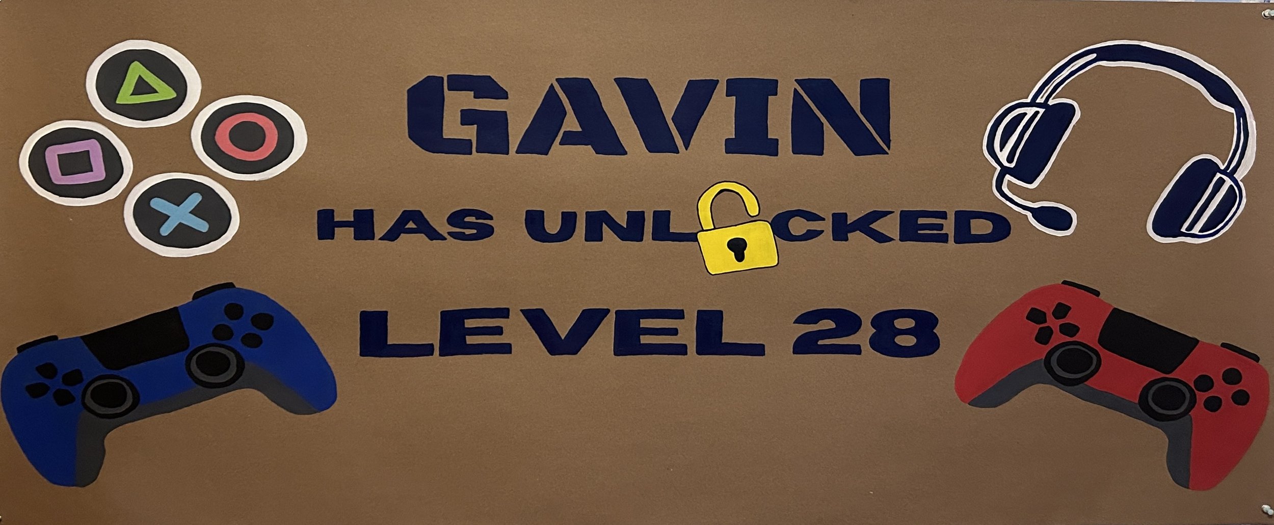 A decorated sign with video game theme elements, including a blue and red game controllers, musical headphones, and a yellow lock icon. The text on the sign reads, 'Gavin has unlocked Level 28'.