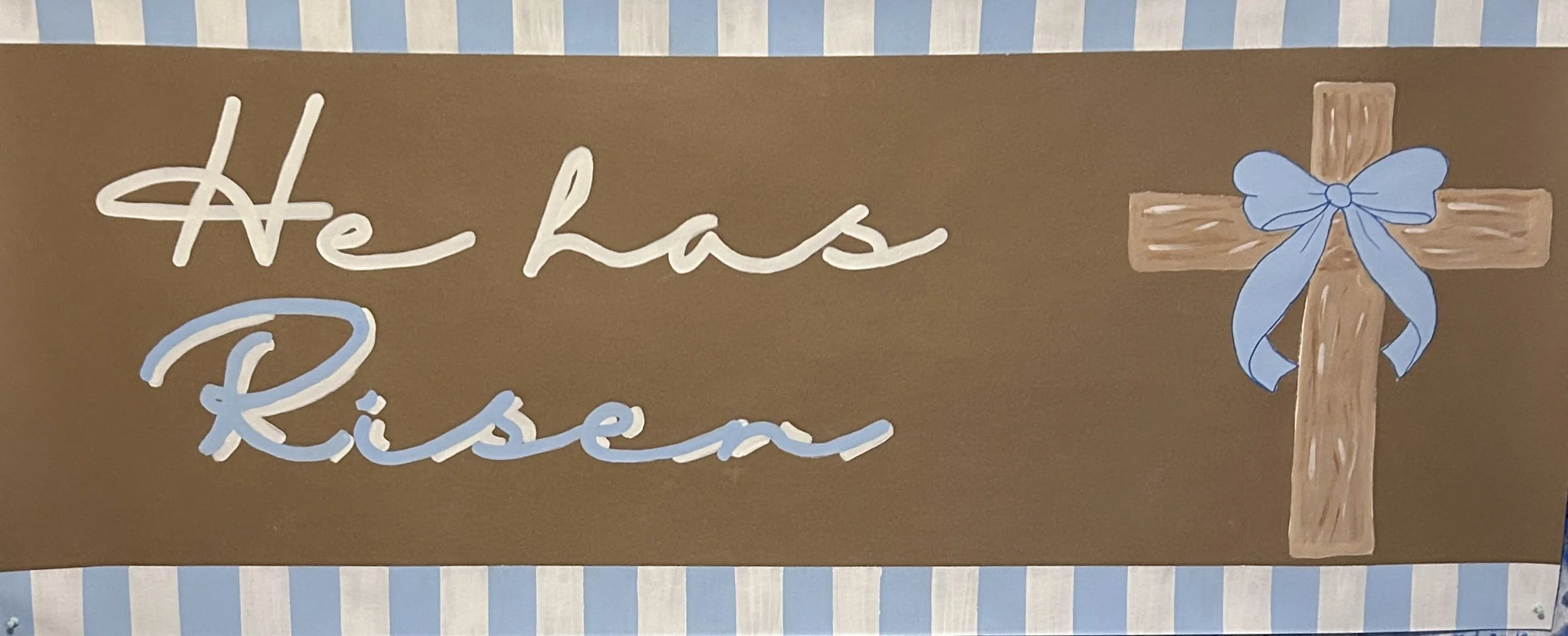 Decorative sign with brown background and blue-striped border, featuring the words 'He has risen' in white and light blue cursive letters, and a wooden cross with a blue ribbon tied around it.