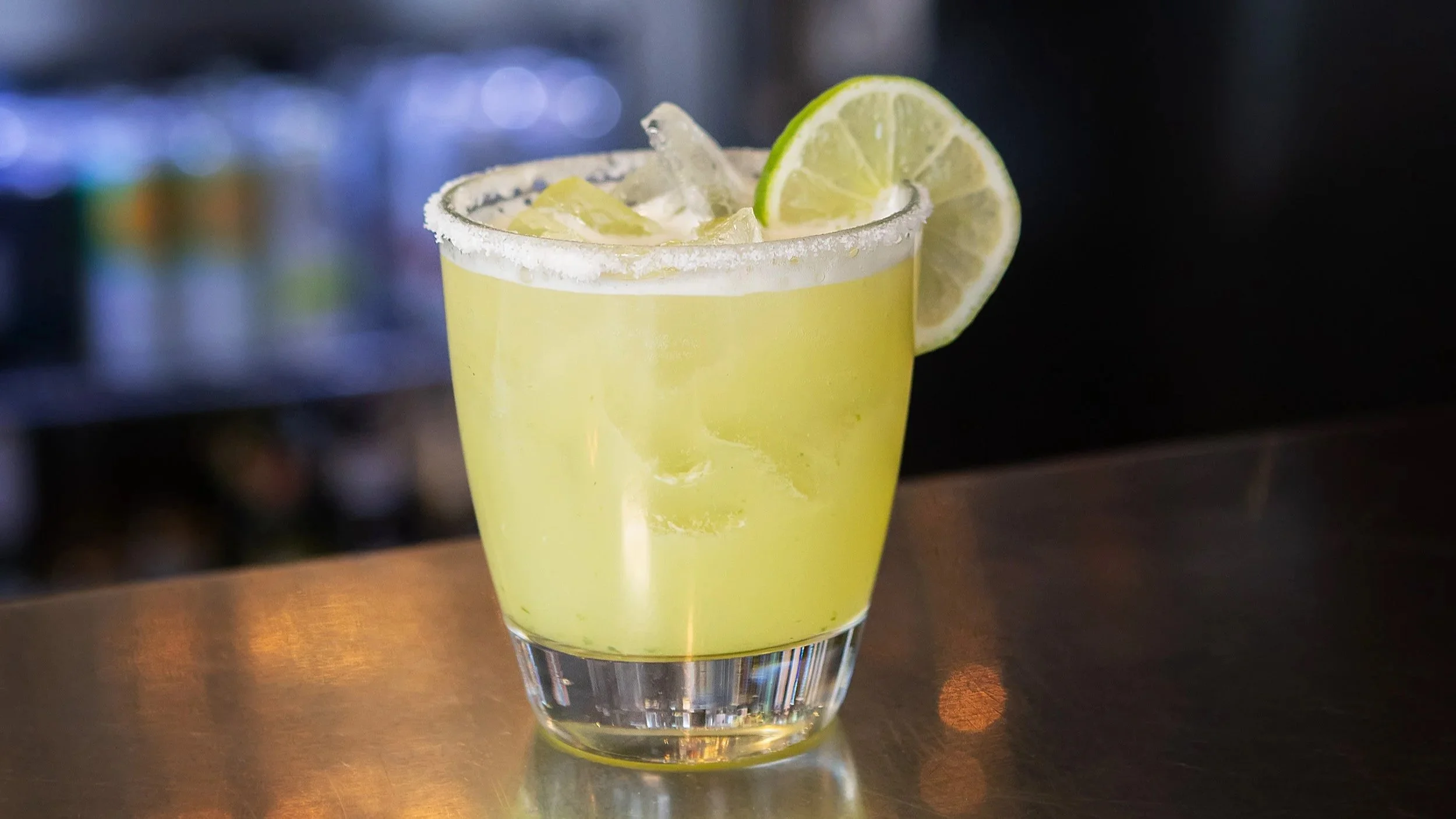 A yellow cocktail garnished with a lime slice and ice cubes in a glass with a sugared rim, sitting on a bar counter.