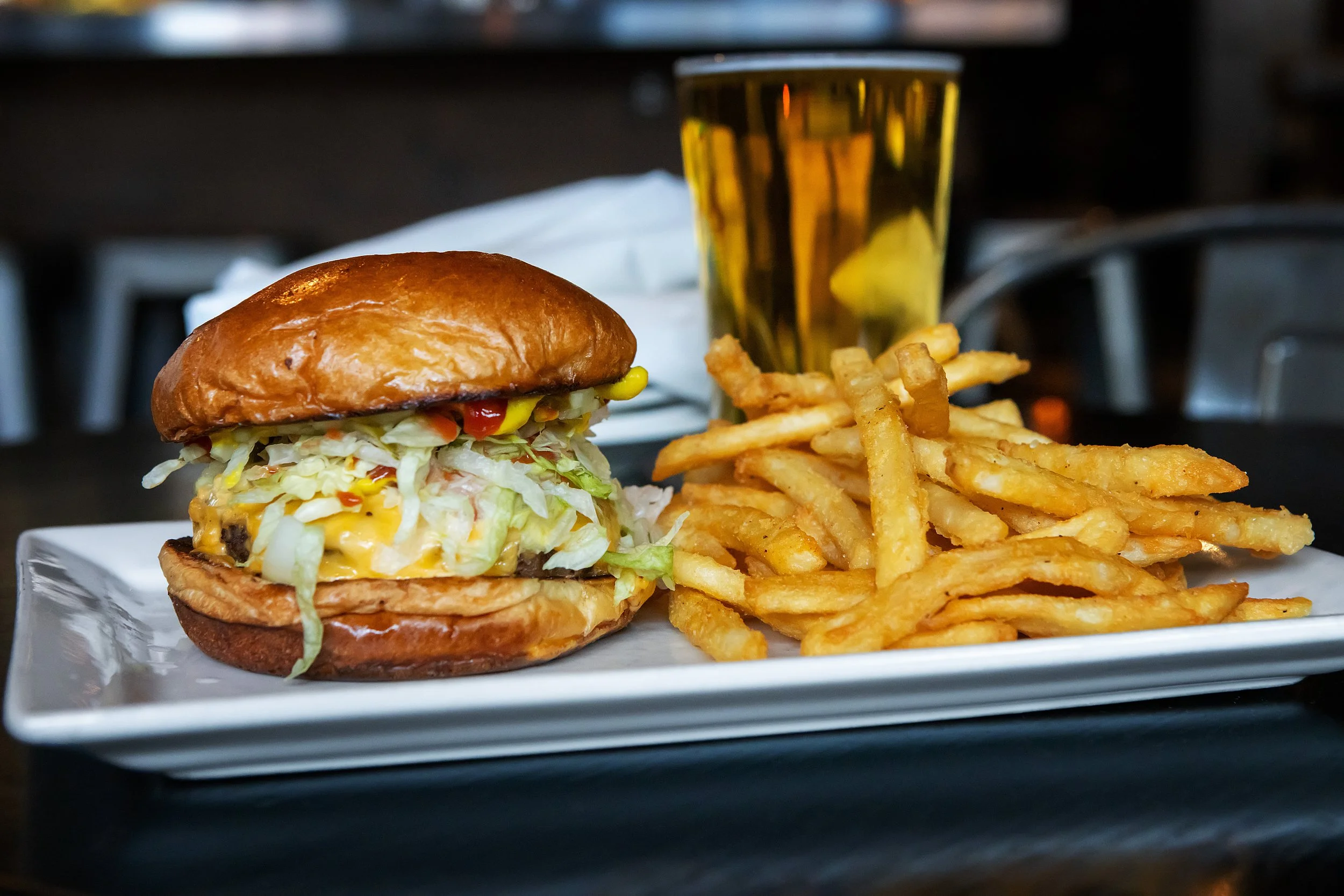 Hamburger with lettuce, tomato, cheese, pickles, and bun, served with French fries and a glass of beer on a white rectangular plate.