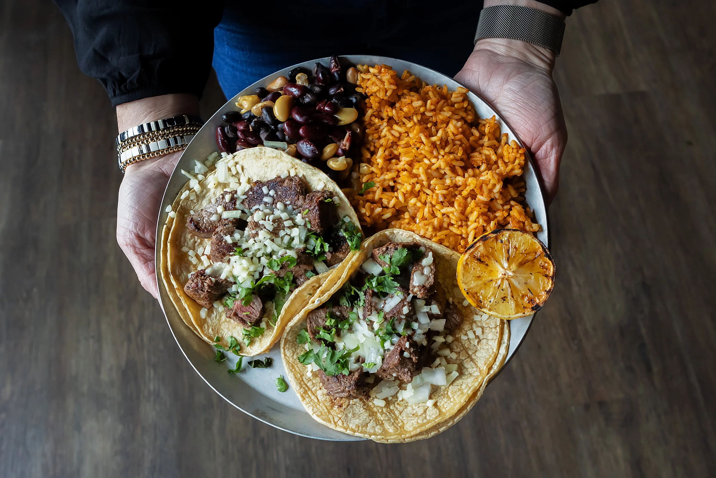 A person holding a tray with three beef tacos, Mexican rice, refried beans, black beans, and a grilled lemon half.