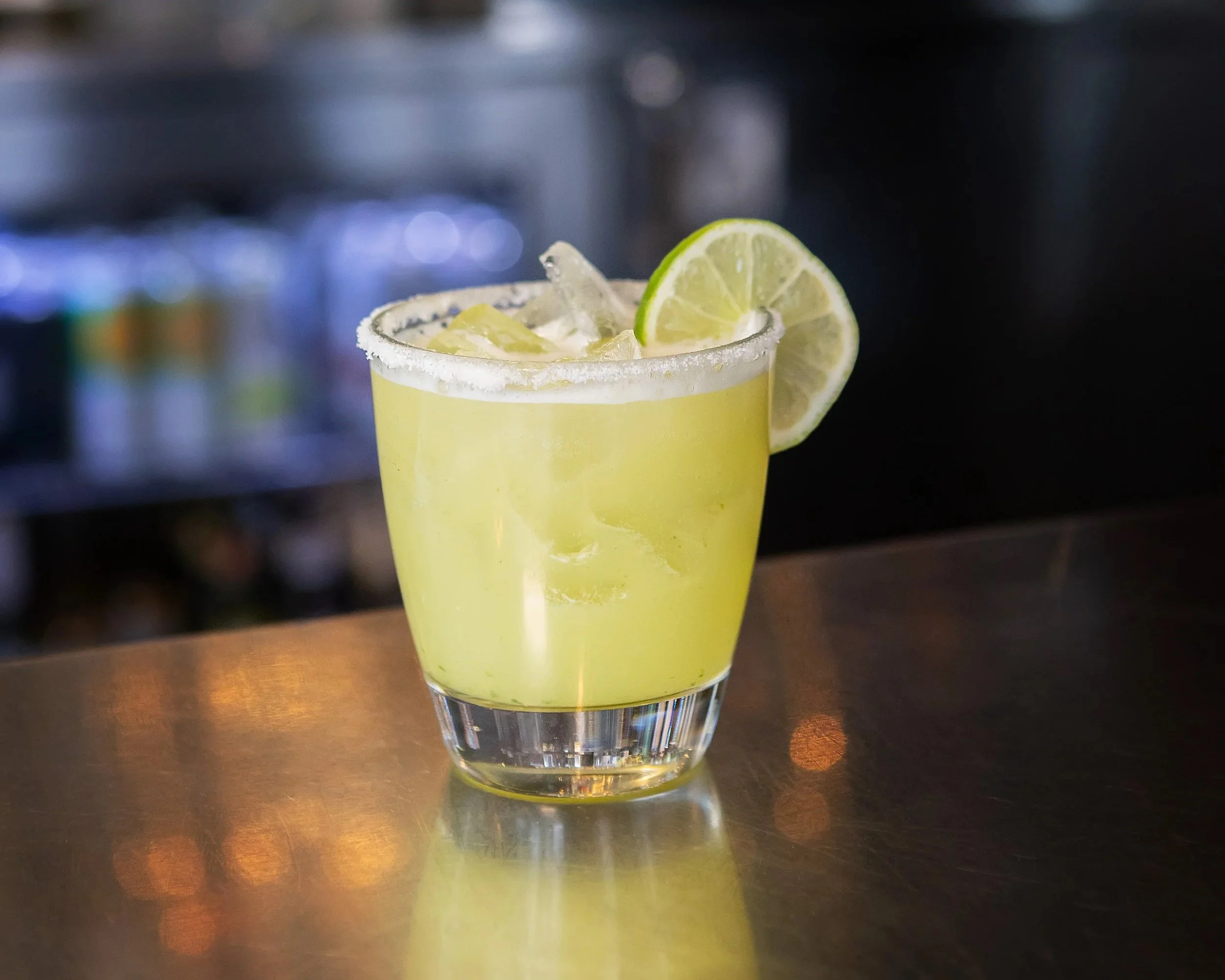 A glass of yellow margarita with lime wedge on the rim, ice cubes, and salt on the glass rim, placed on a dark bar counter.