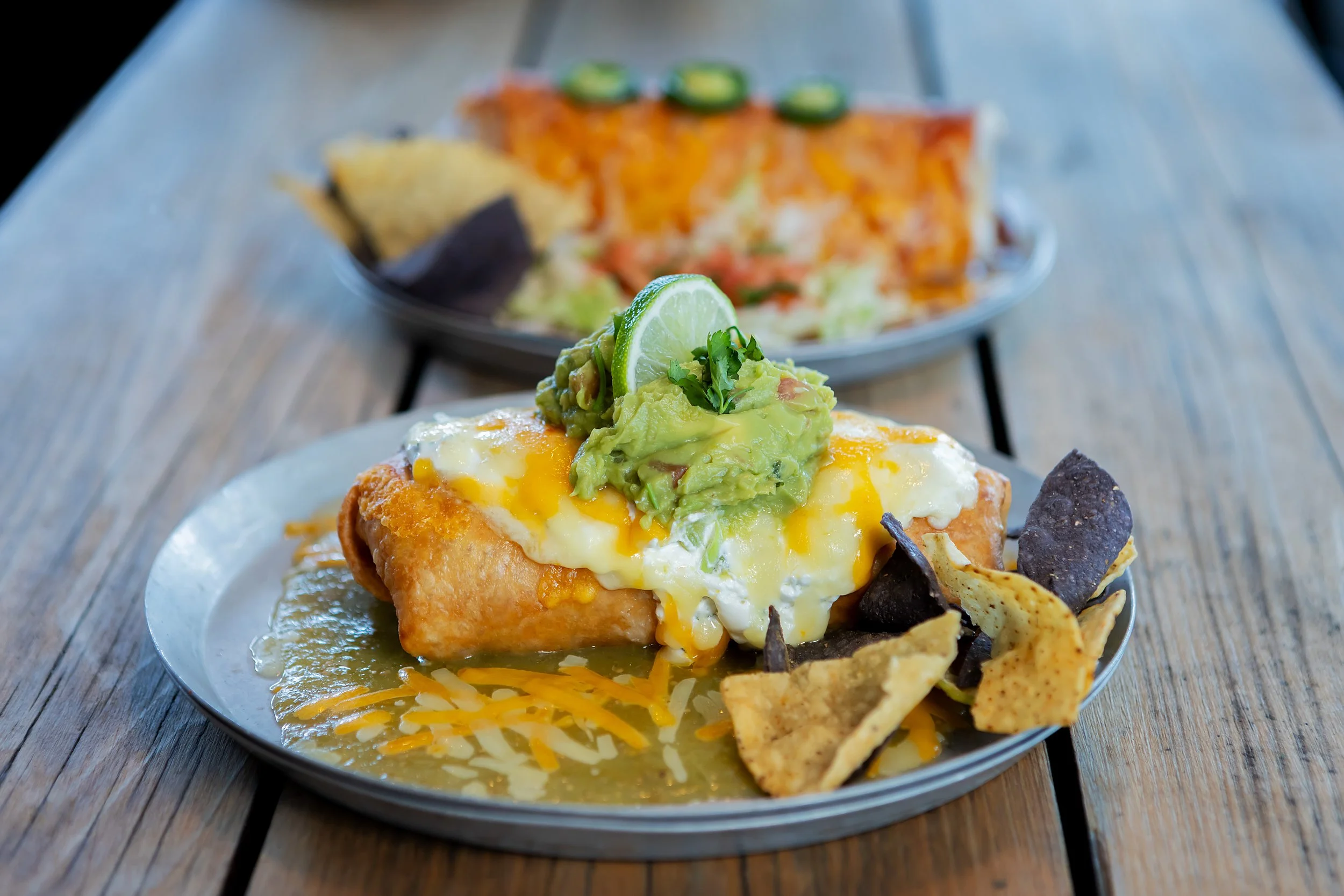 A plate of huevos rancheros with a fried tortilla, topped with eggs, melted cheese, guacamole, a lime wedge, and garnished with cilantro, with blue corn chips around the plate. In the background, a dish of nachos with jalapeños and melted cheese.